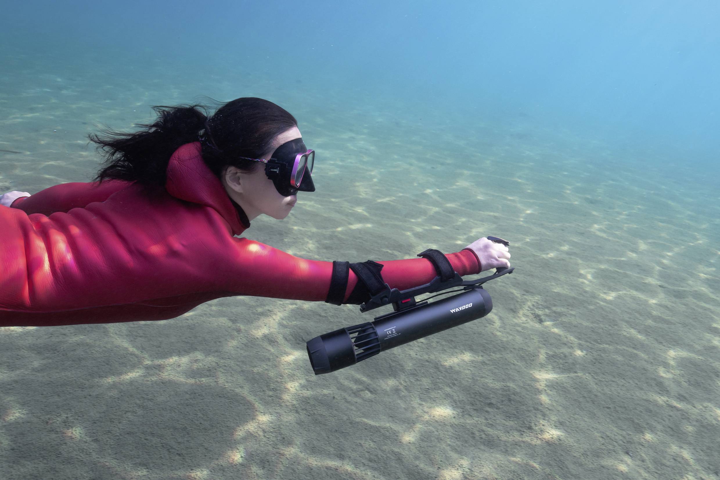 Una persona in muta rossa e maschera nuota sott'acqua con un piccolo scooter subacqueo sopra un fondale marino sabbioso.