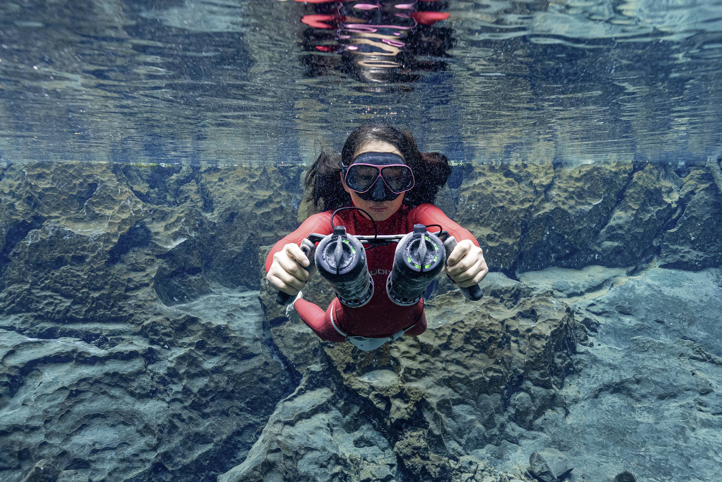 Una persona è immersa sott'acqua in un ambiente cristallino, con due dispositivi, probabilmente fotocamere subacquee, tra le mani.