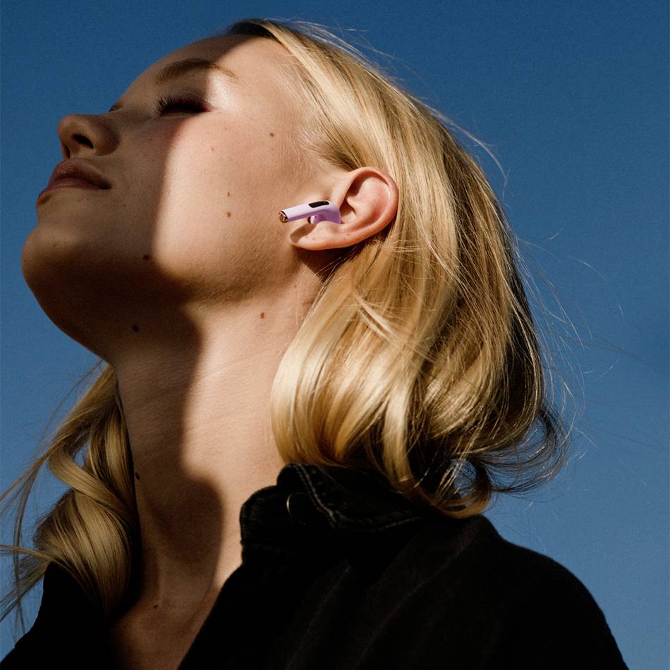 Una persona dai capelli biondi guarda verso il sole con gli occhi chiusi. Indossa auricolari wireless. Il cielo è limpido.