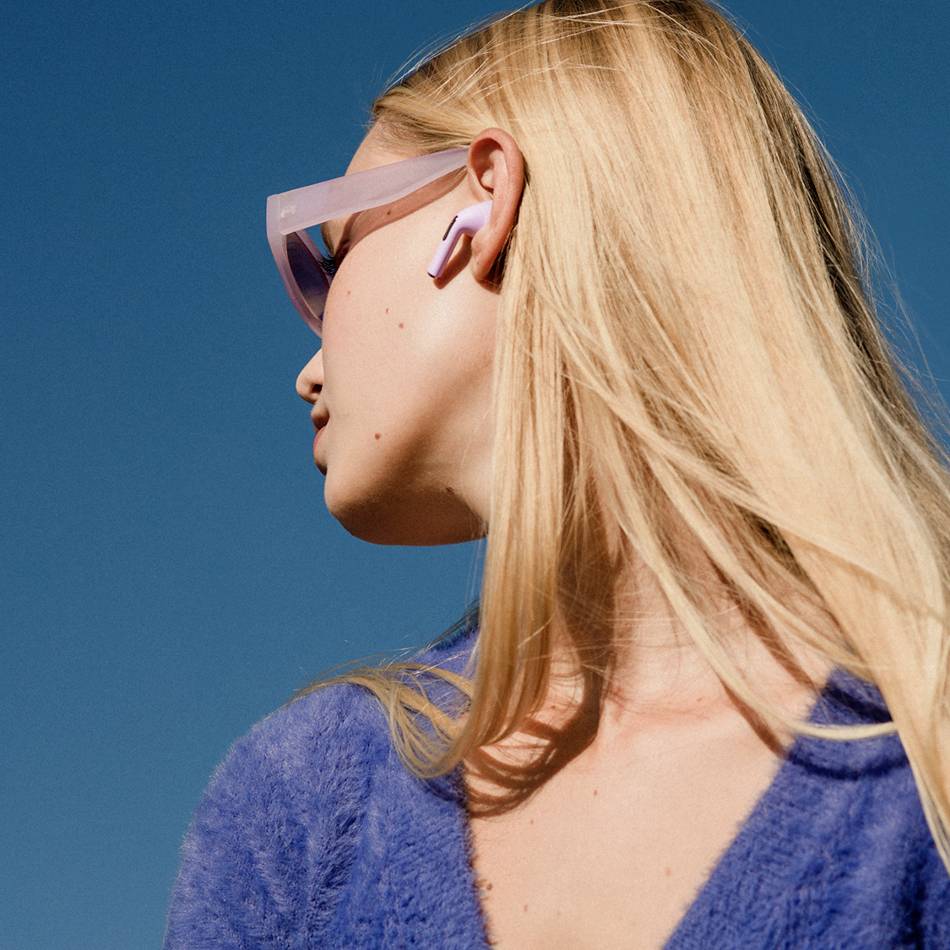 Donna con capelli biondi indossa occhiali da sole blu e cardigan blu, guarda verso il cielo. Ha auricolari wireless nell'orecchio.