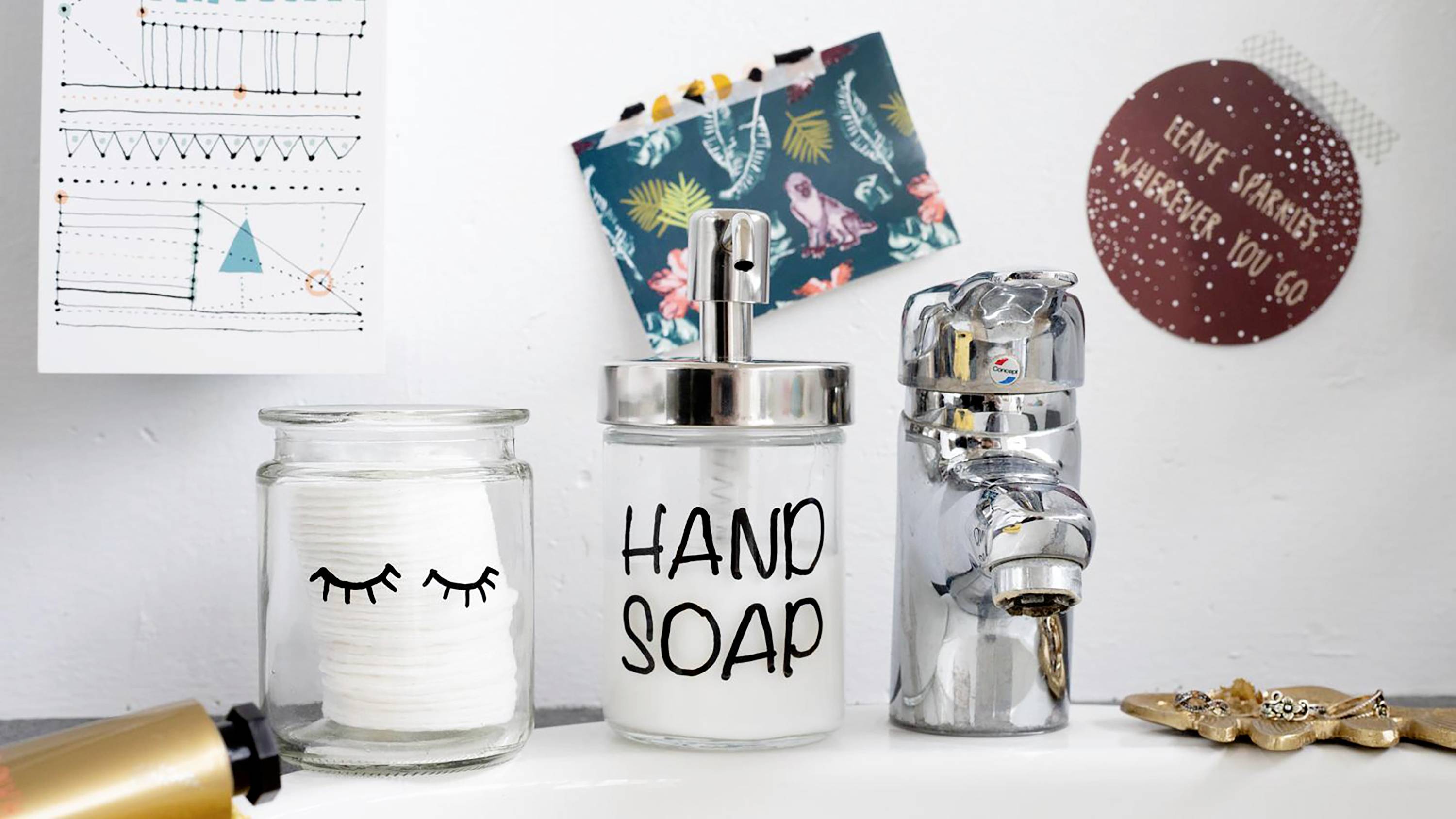 Bicchieri con dischetti di cotone, sapone liquido in un dispenser con la scritta 'Hand Soap' e elastici per capelli su uno scaffale del bagno.