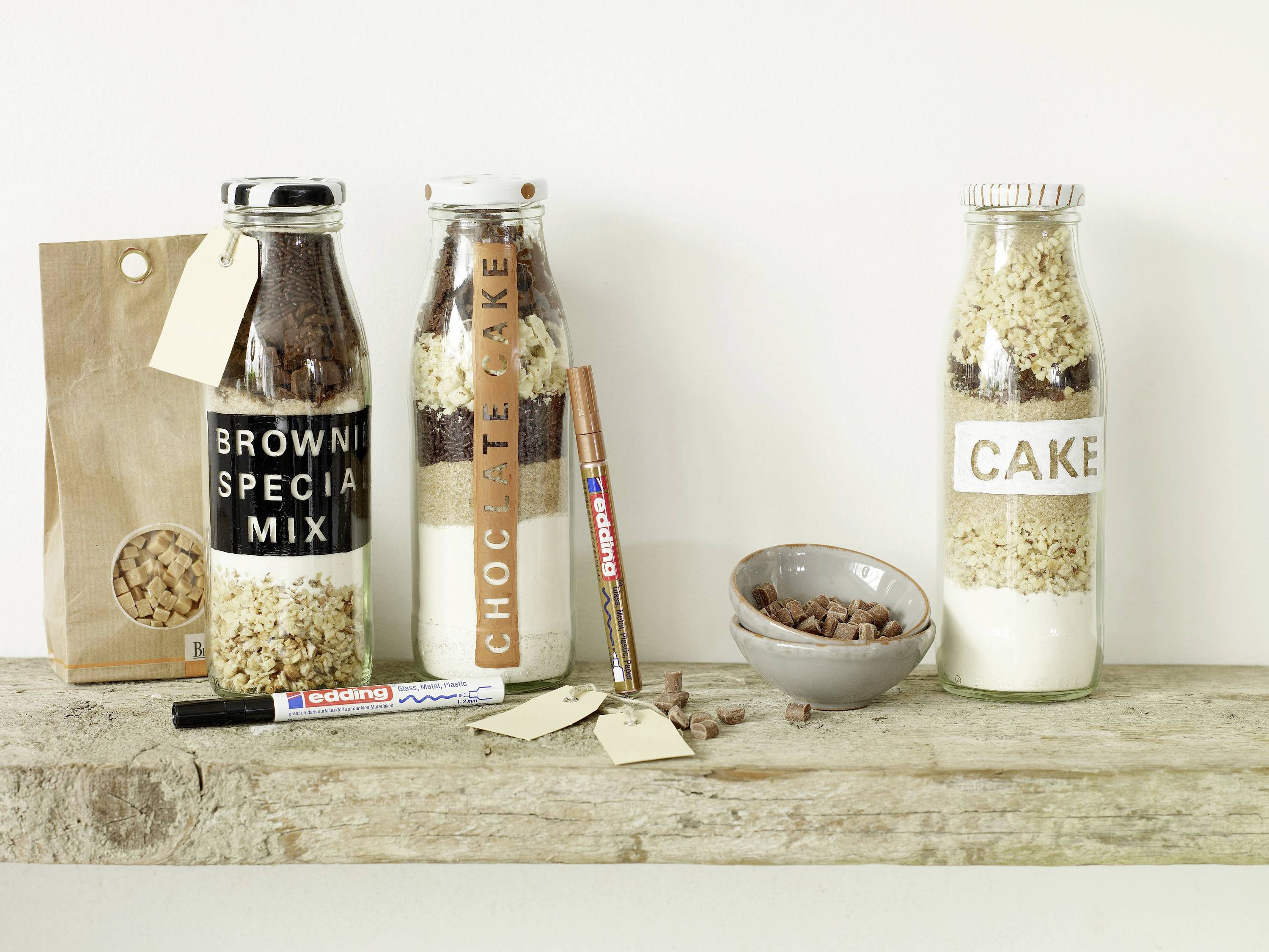 'Tre diversi mix per dolci in bottiglie su un tagliere di legno, accanto a una ciotola con fave di cacao e vari materiali per l'etichettatura.'