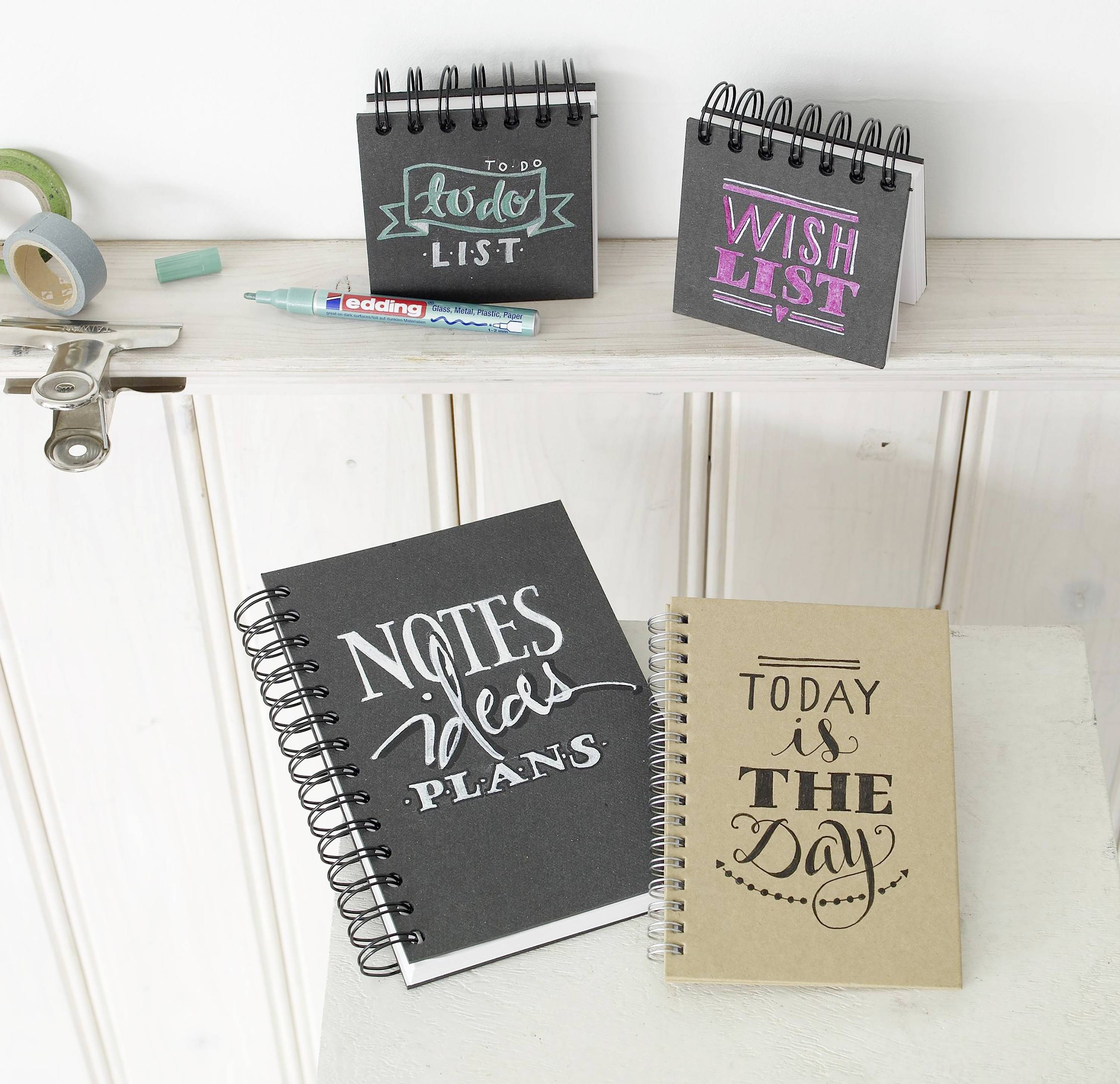 Cinque quaderni su un supporto di legno. I titoli recitano: 'Lista delle cose da fare', 'Lista dei desideri', 'Piani - Note - Idee' e 'Oggi è il giorno'. Accessori per la scrittura sono posizionati accanto.