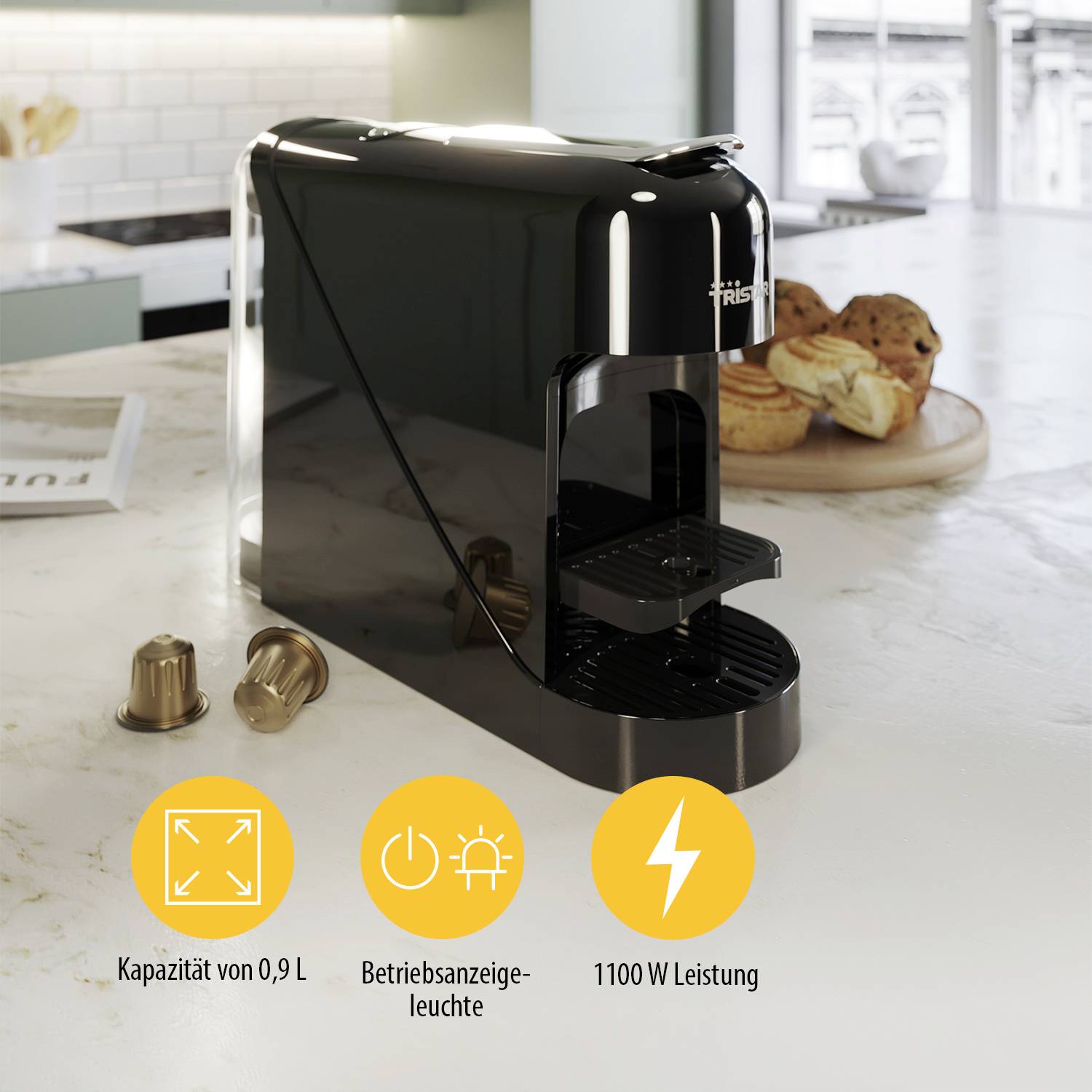 Una macchina da caffè a capsule nera su un piano da cucina. Accanto ci sono due capsule e dolcetti. Testo: 'Capacità di 0,9 L', 'Indicatore di funzionamento', 'Potenza da 1100 W'.