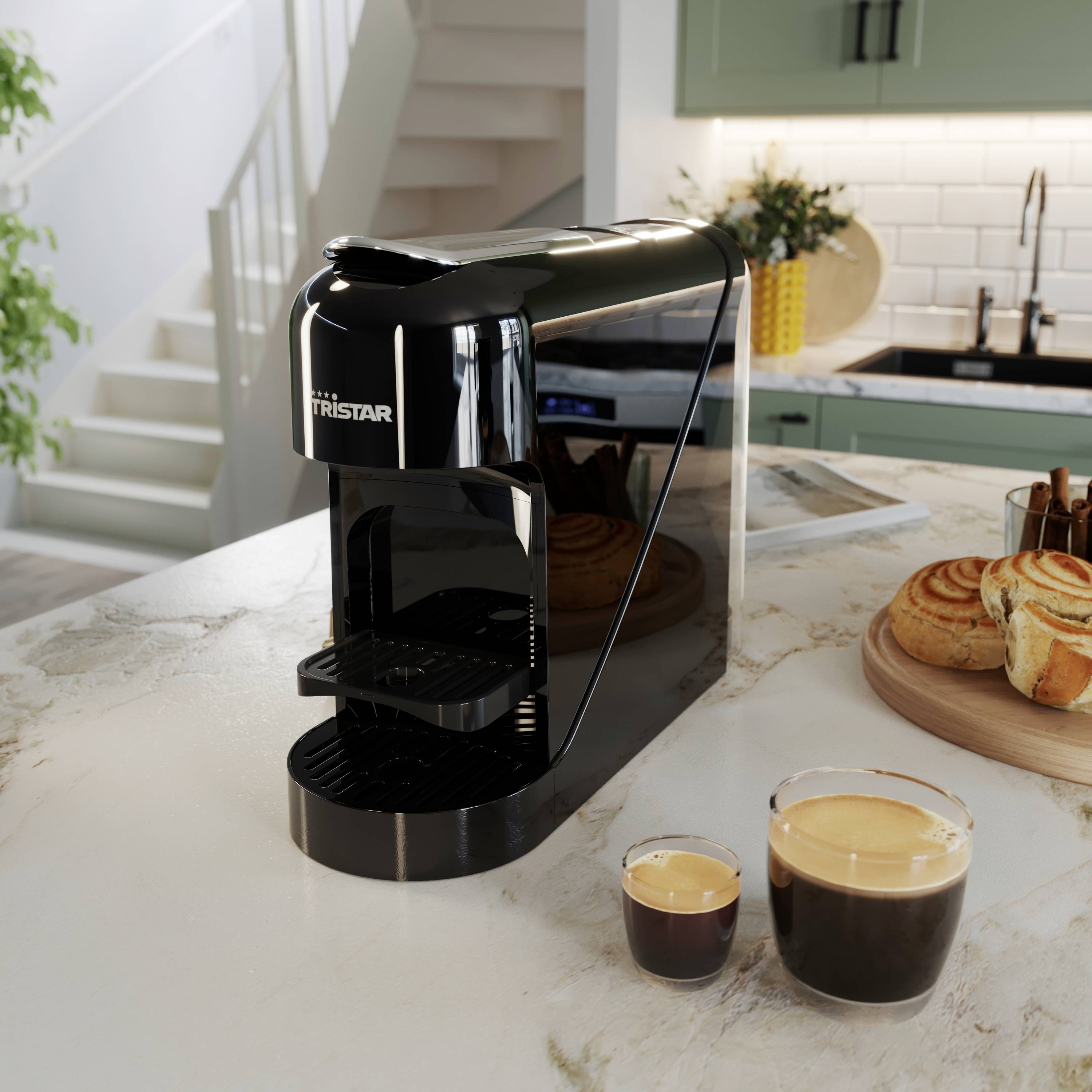 Una macchina da caffè nera è posizionata su un piano di lavoro della cucina accanto a due tazze piene e alcuni dolci. Sullo sfondo sono visibili una scala e un lavandino.