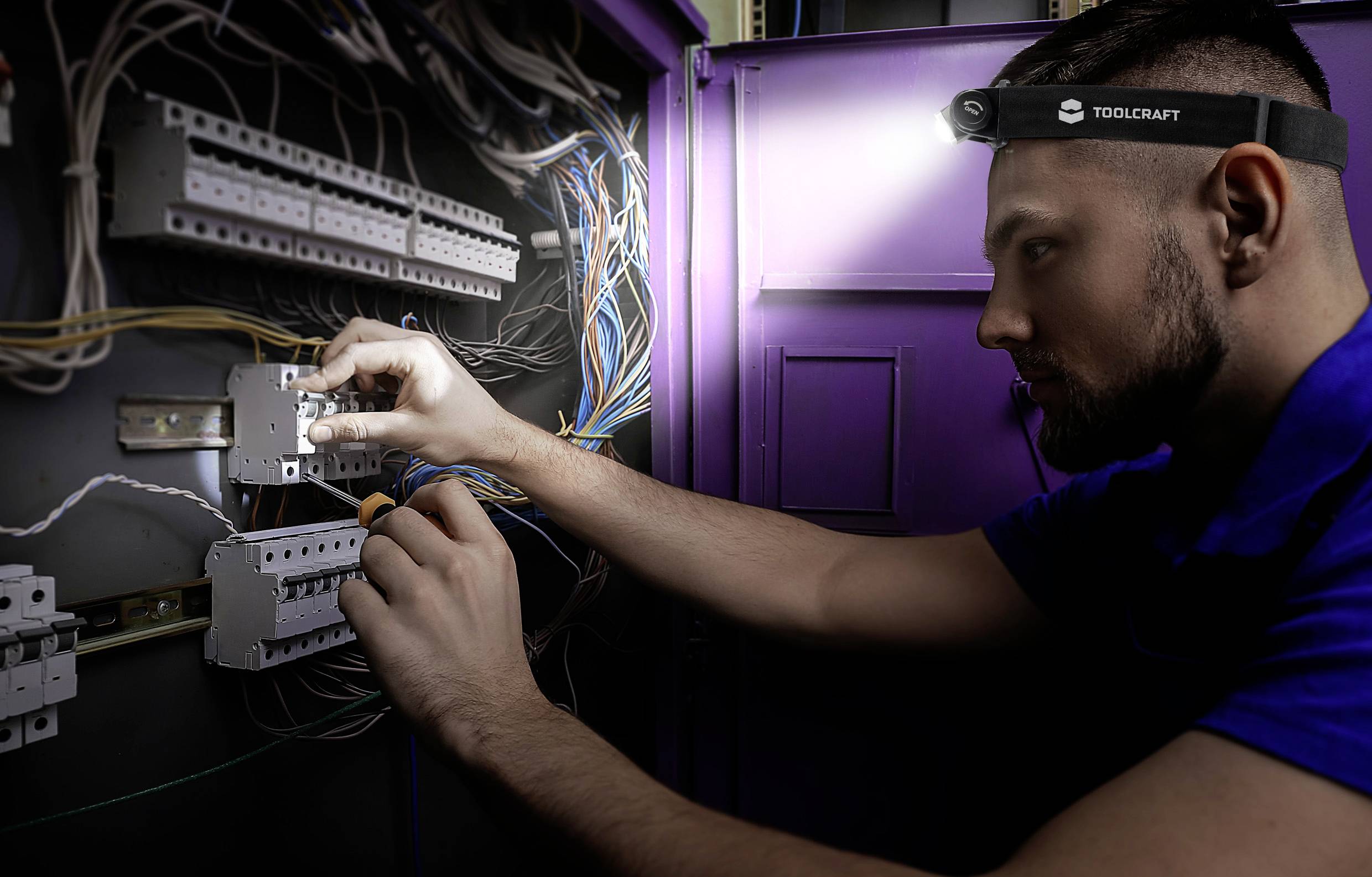 Un tecnico lavora con una lampada frontale su un quadro elettrico, collegando cavi a una scheda. Lavori elettrici in corso.