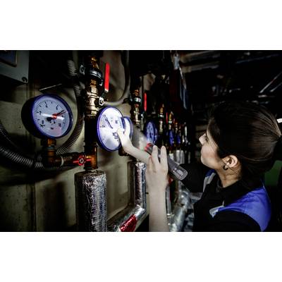 Una persona controlla diversi indicatori di pressione su tubazioni e valvole industriali con una torcia elettrica.