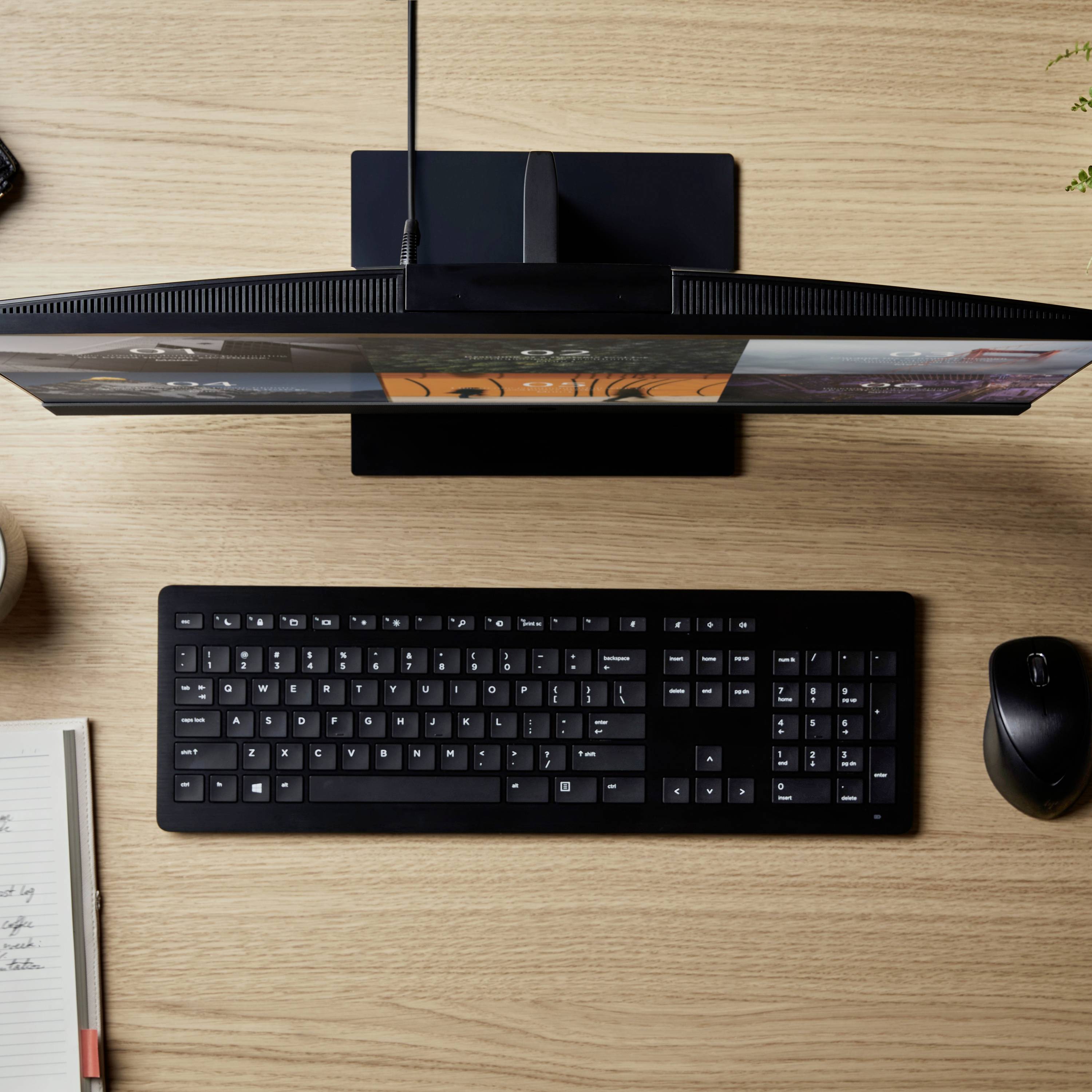 Un posto di lavoro con computer, composto da un monitor, una tastiera e un mouse su un tavolo di legno. Accanto sono posizionati un quaderno e una penna.