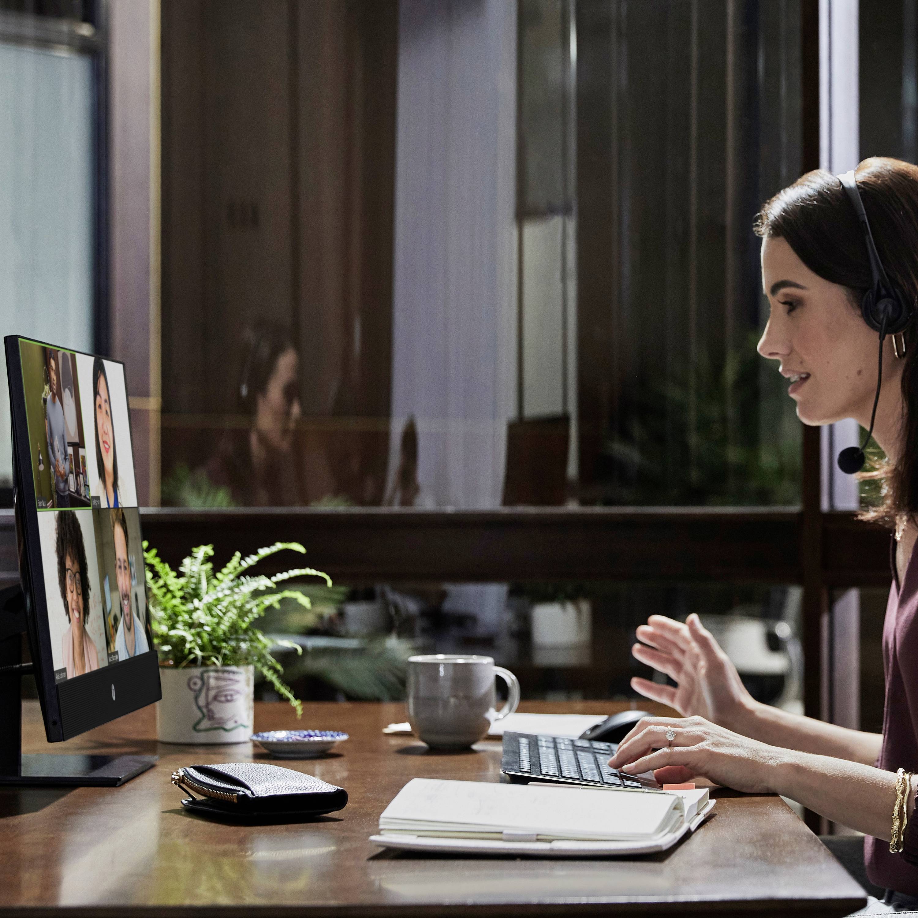 Una donna con un headset in un ufficio sta tenendo una videoconferenza con quattro persone su un monitor. Scrivania con laptop e quaderno.