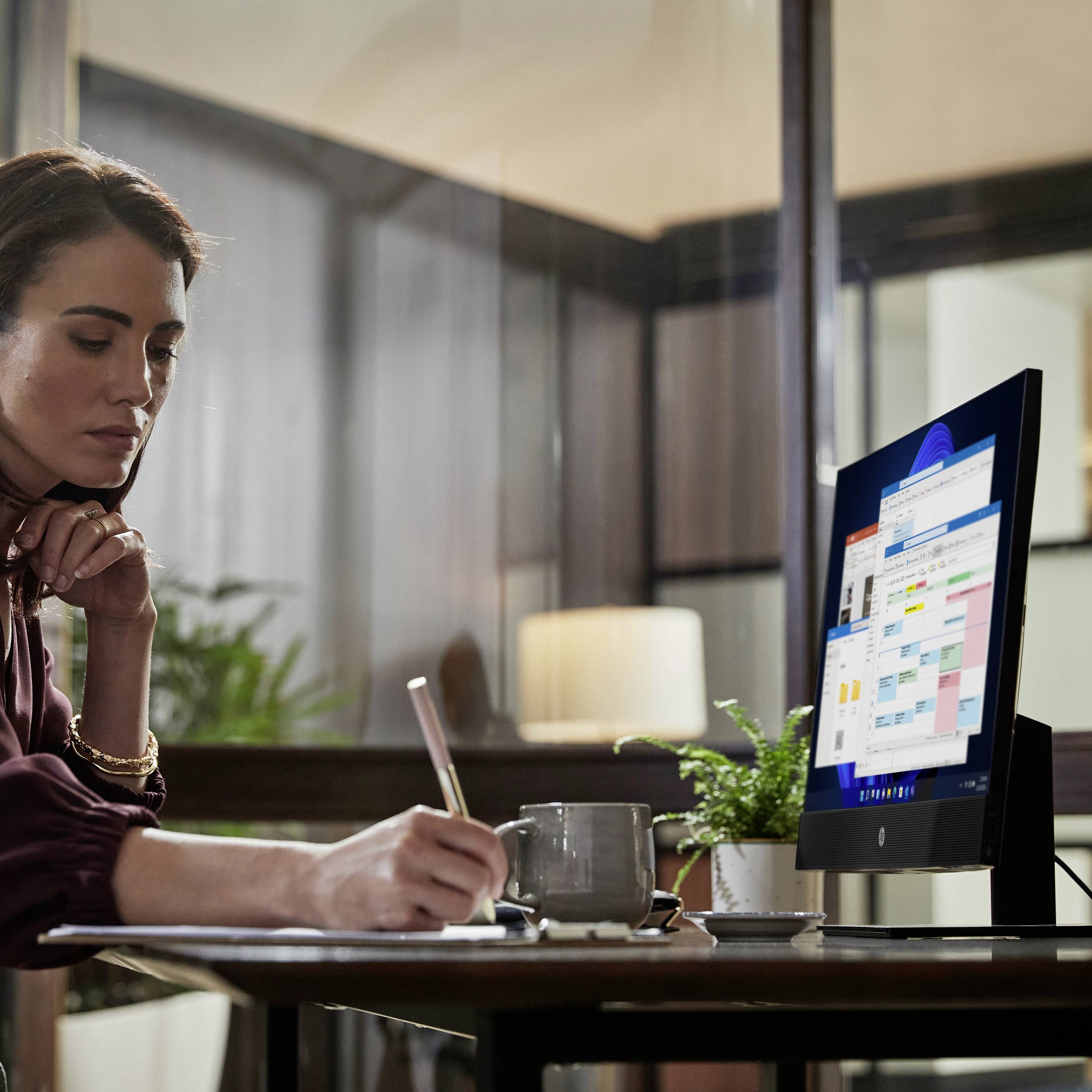 Una donna è seduta a una scrivania in un ufficio moderno, scrive con una penna su un foglio di carta mentre guarda uno schermo del computer.