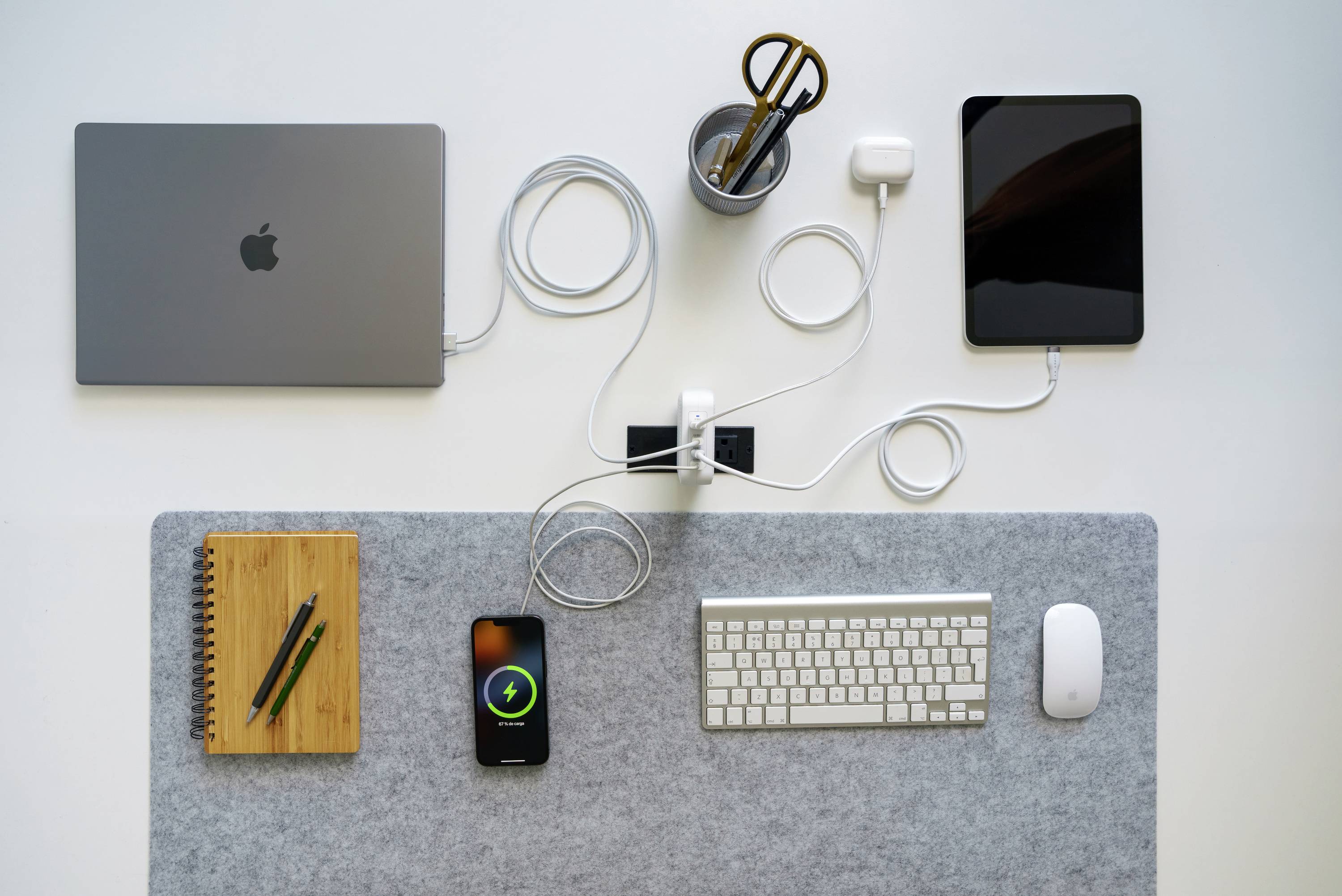 Una scrivania ordinata con laptop, tablet, tastiera, mouse, quaderno, penne e un cavo di ricarica. Uno smartphone è in carica.