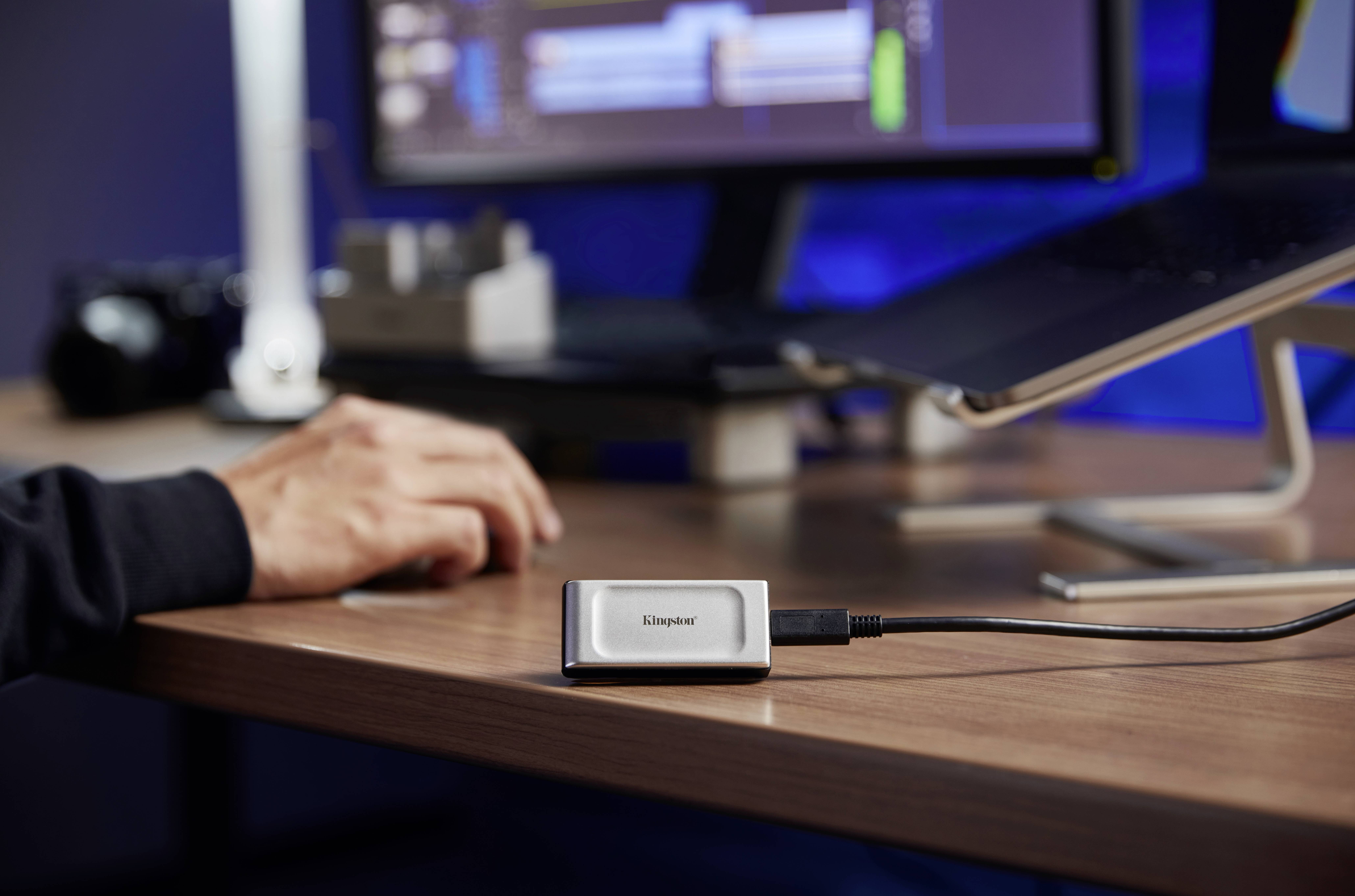 Sul tavolo, un hard disk esterno è posizionato accanto a un monitor e a una mano poggiata sul mouse del computer. Sullo sfondo, è in esecuzione un software.