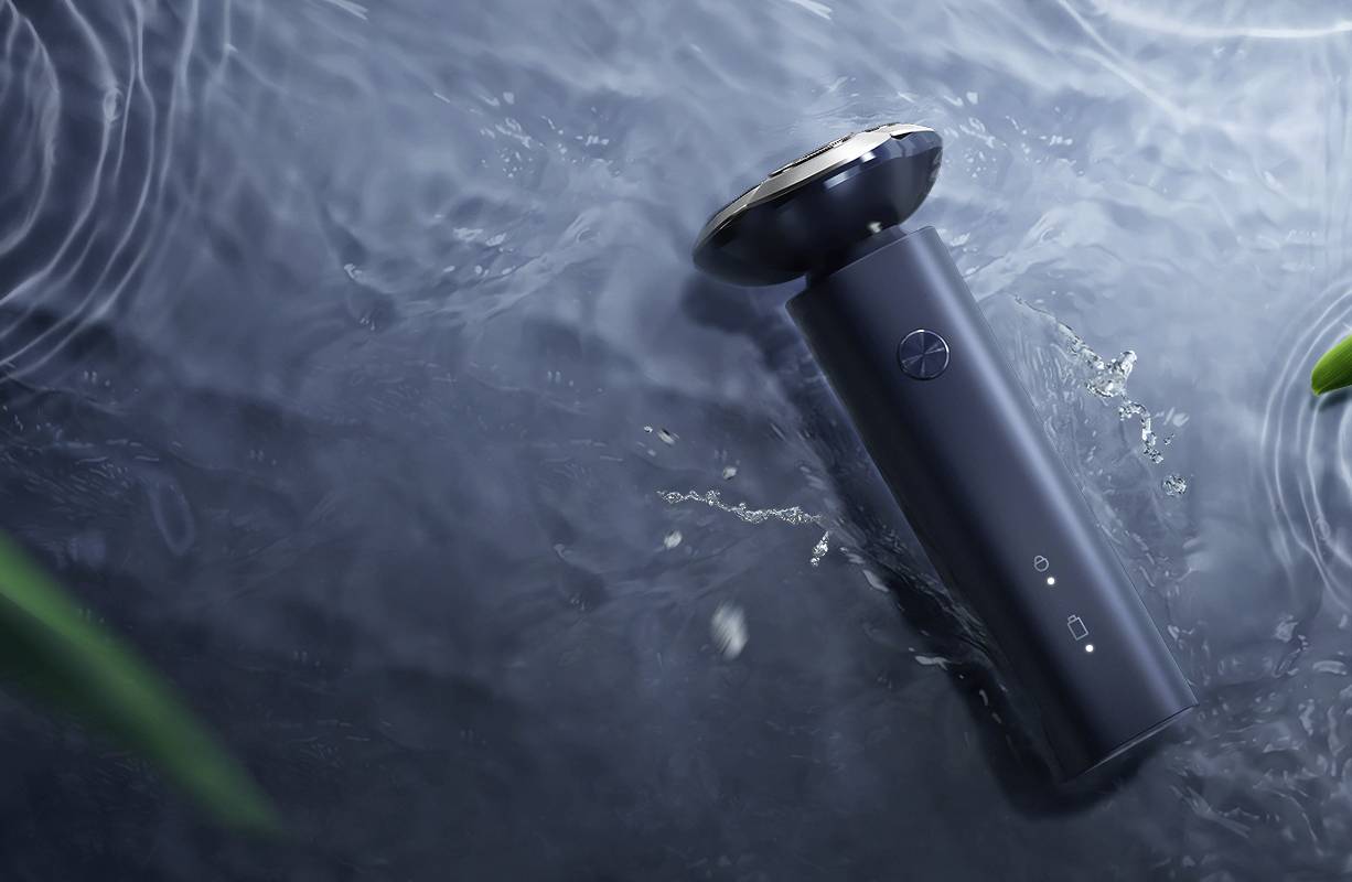 Un rasoio elettrico galleggia nell'acqua. Involucro scuro con testa argentata. Ambiente con superficie acquatica ed elementi vegetali.
