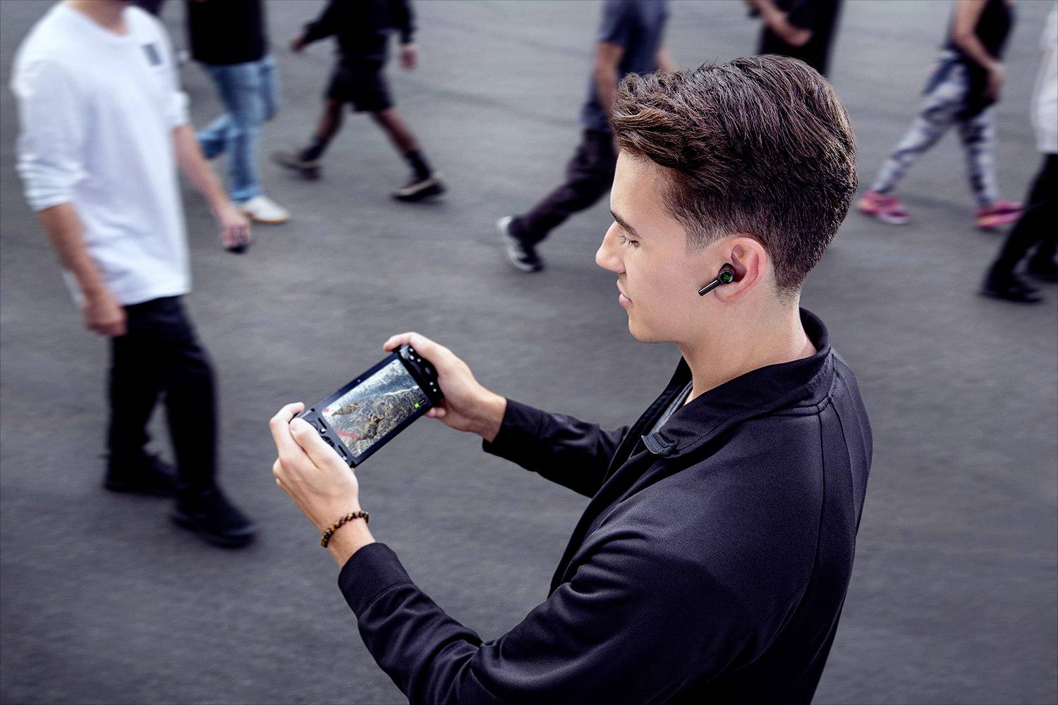 Un giovane uomo gioca concentrato a un gioco su un dispositivo portatile in un ambiente affollato, indossando auricolari wireless.