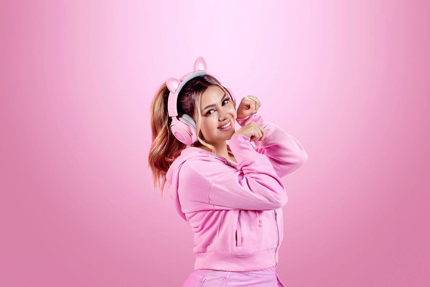 Una persona vestita di rosa indossa delle cuffie con un design a forma di gatto. Sorride e posa davanti a uno sfondo rosa.