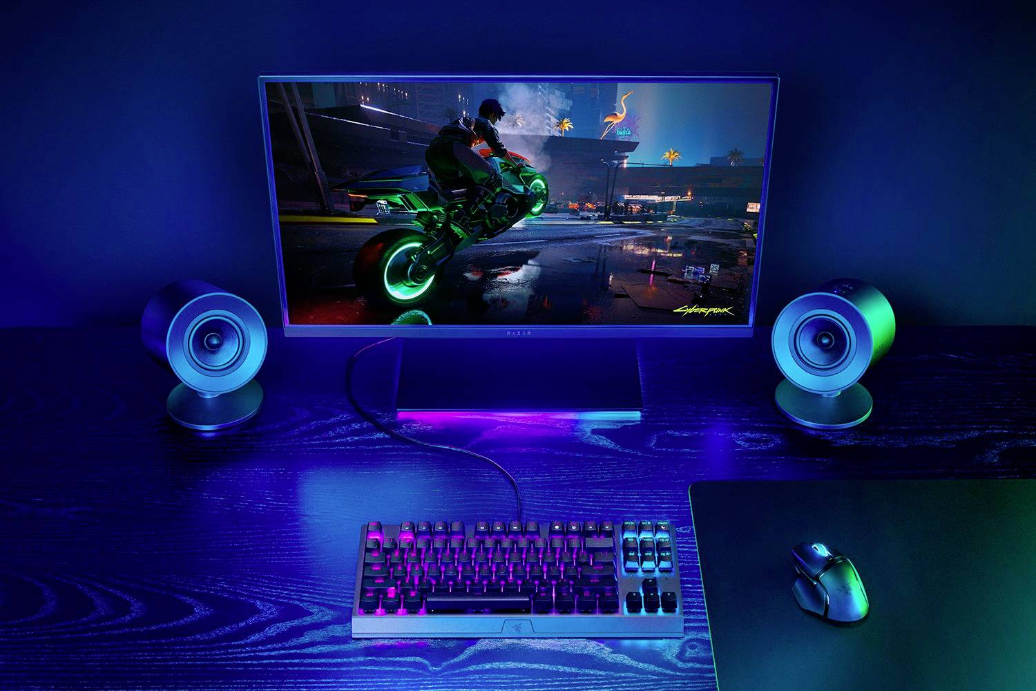 Un monitor del computer mostra un videogioco con un motociclista in una città futuristica, circondato da tastiera e altoparlanti.