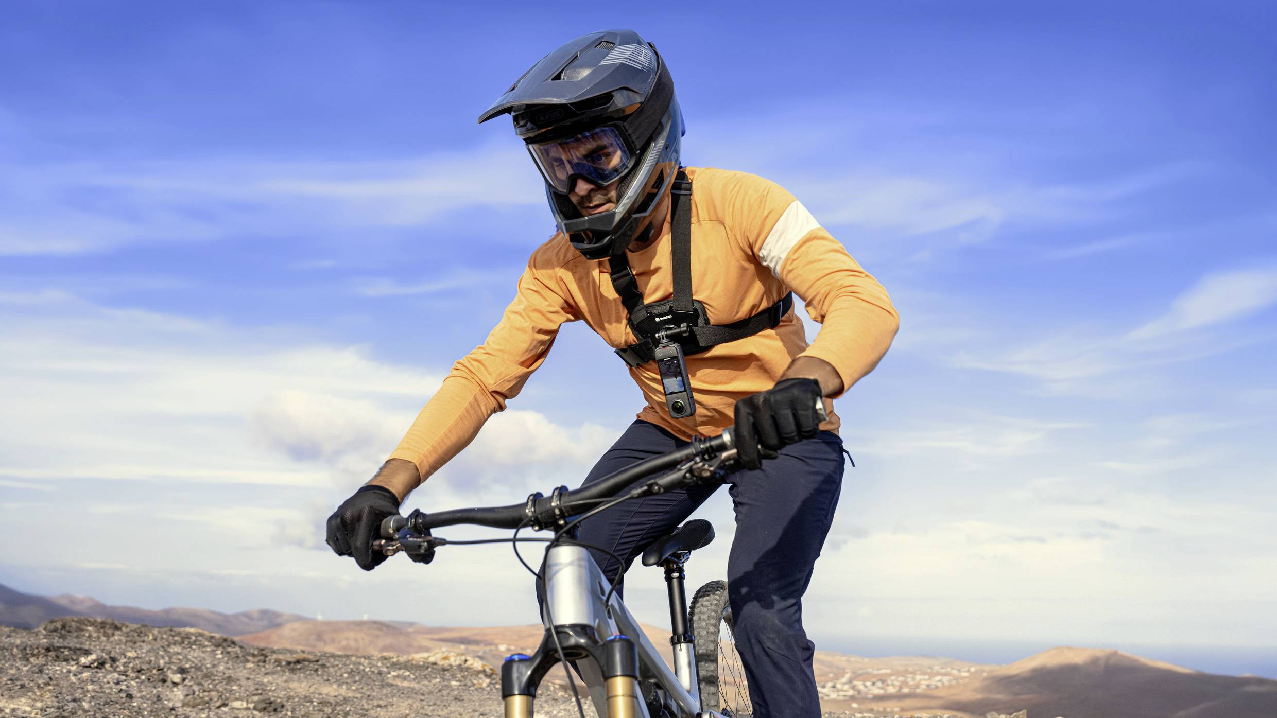 Un uomo con abbigliamento protettivo guida una mountain bike su una collina con un cielo limpido. Lo sfondo mostra un paesaggio vasto e aperto.