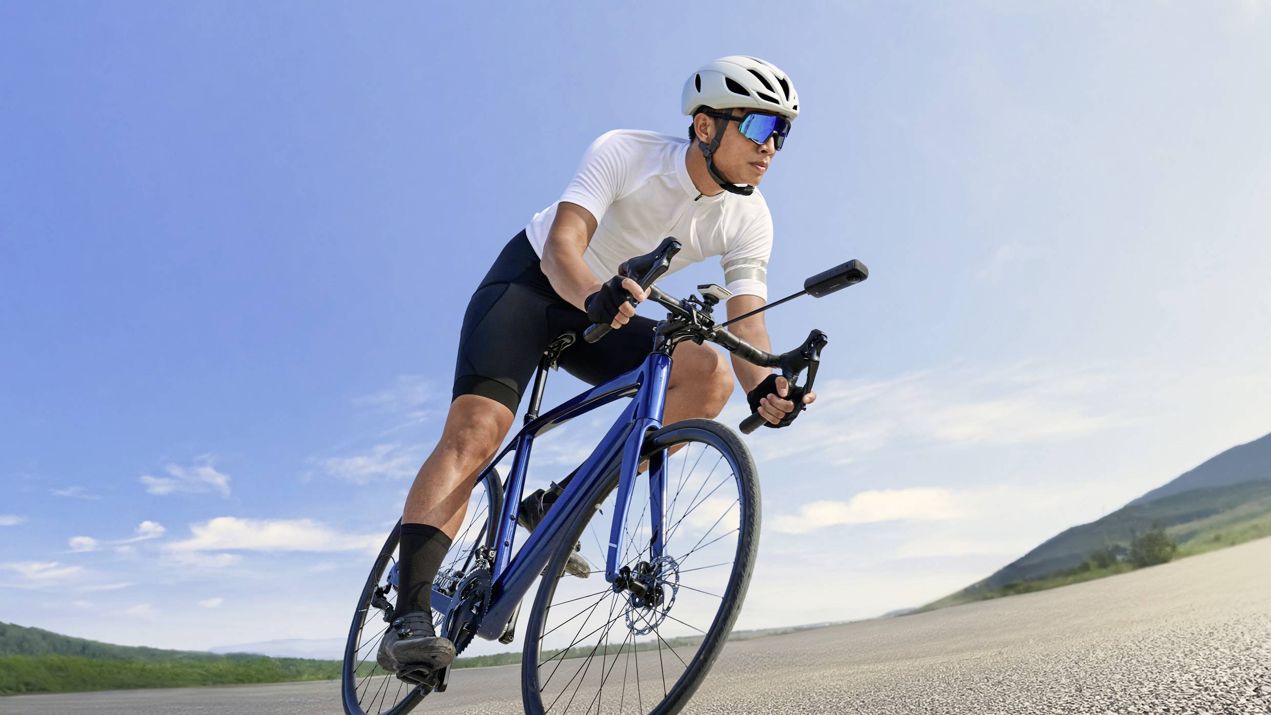 Un ciclista in abbigliamento sportivo pedala su una strada statale, circondato da un cielo blu e un paesaggio verde, con il sole che splende.