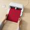 Una persona tiene un foglio rosso in un plotter da taglio su un tavolo di legno, pronta per il taglio. Dispositivo in primo piano, accessori a sinistra.