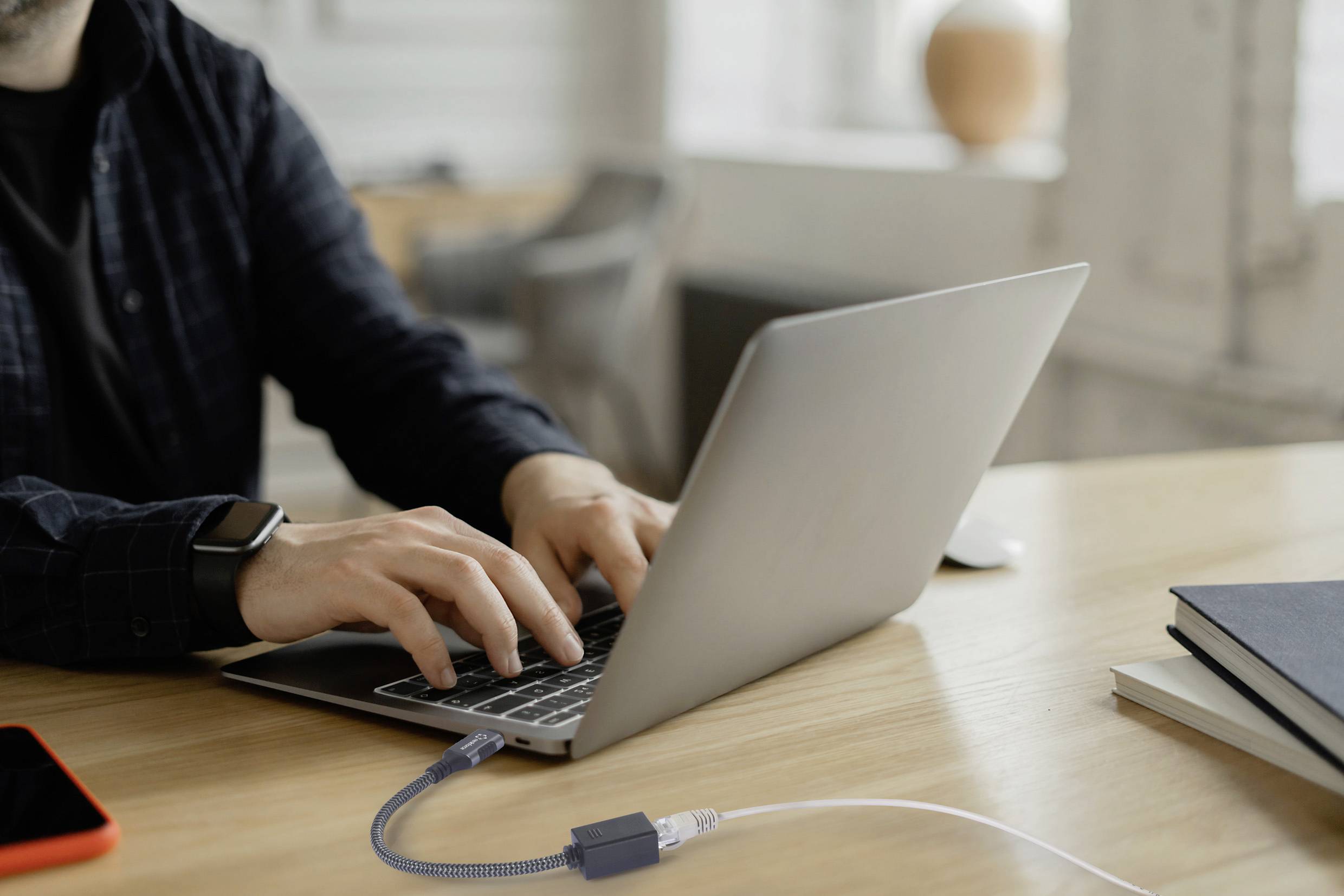 Una persona sta lavorando su un laptop posto su un tavolo di legno, circondato da libri e un adattatore di rete. In primo piano, uno smartphone con una custodia rossa.