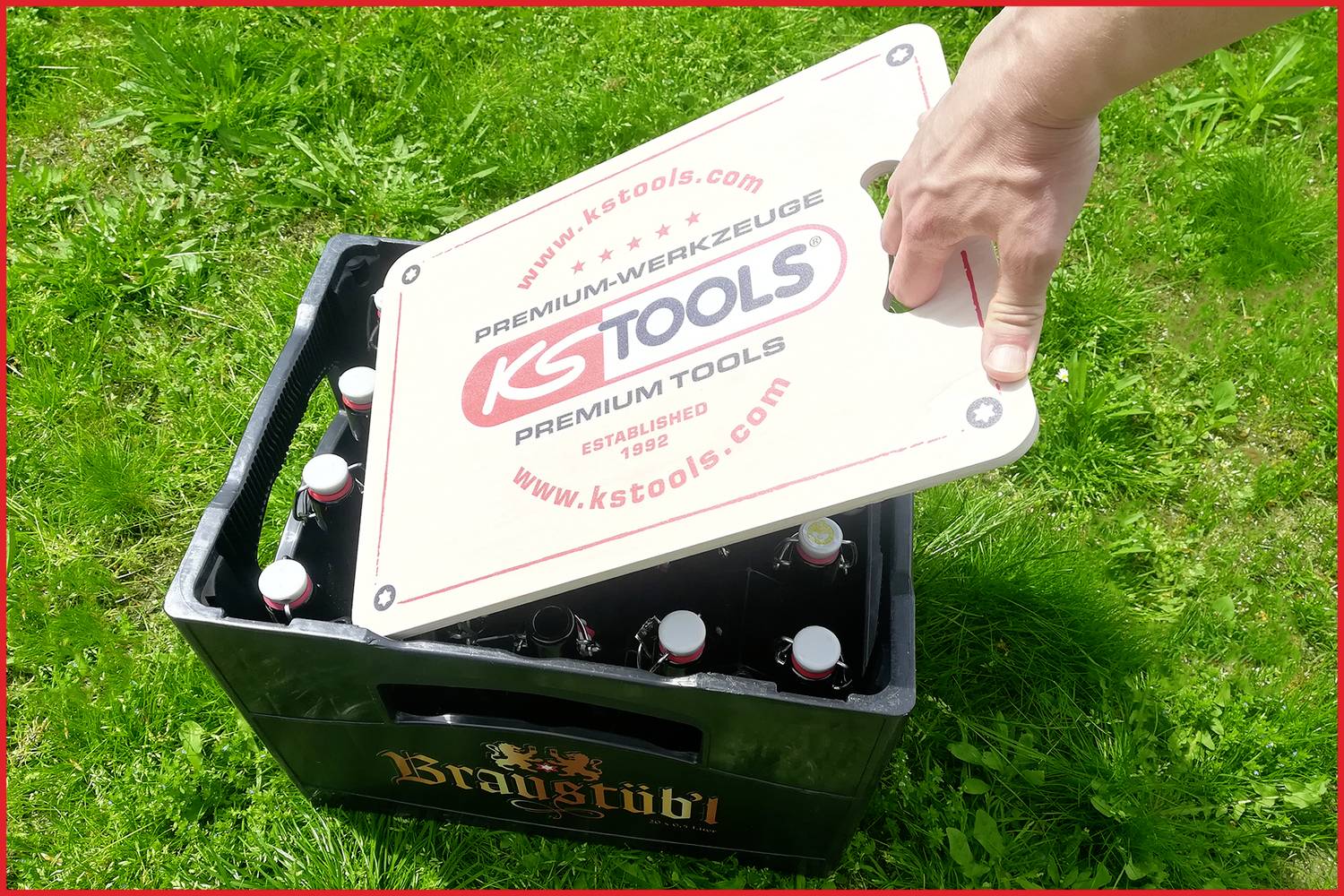 Una mano solleva un apribottiglie stampato da una cassa di bevande su un prato. Sull'apribottiglie è scritto 'KS Tools'.