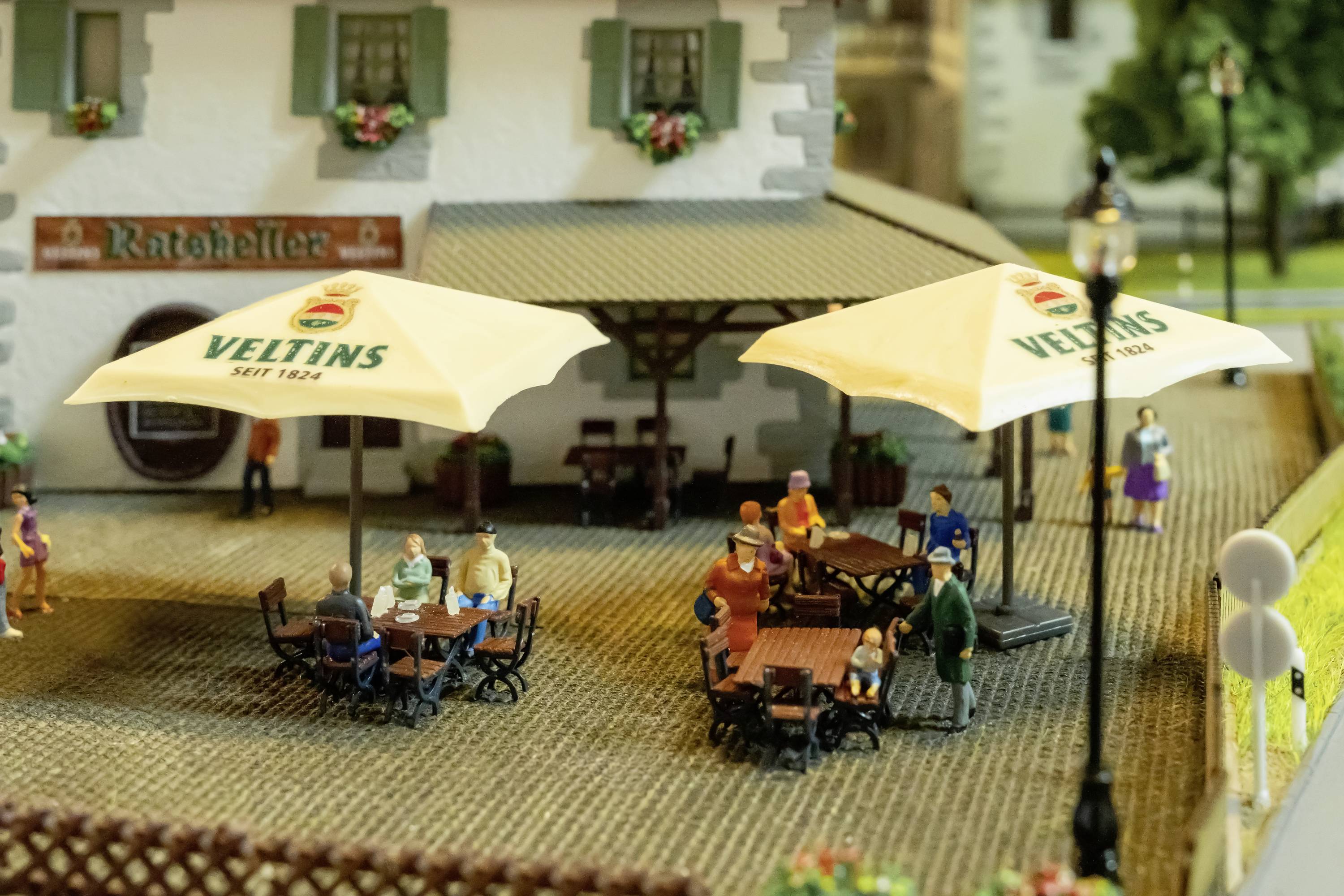 Scena in miniatura di una strada con persone sedute a tavoli sotto ombrelloni all'aperto, con un edificio sullo sfondo che reca la scritta 'Ratskeller'.