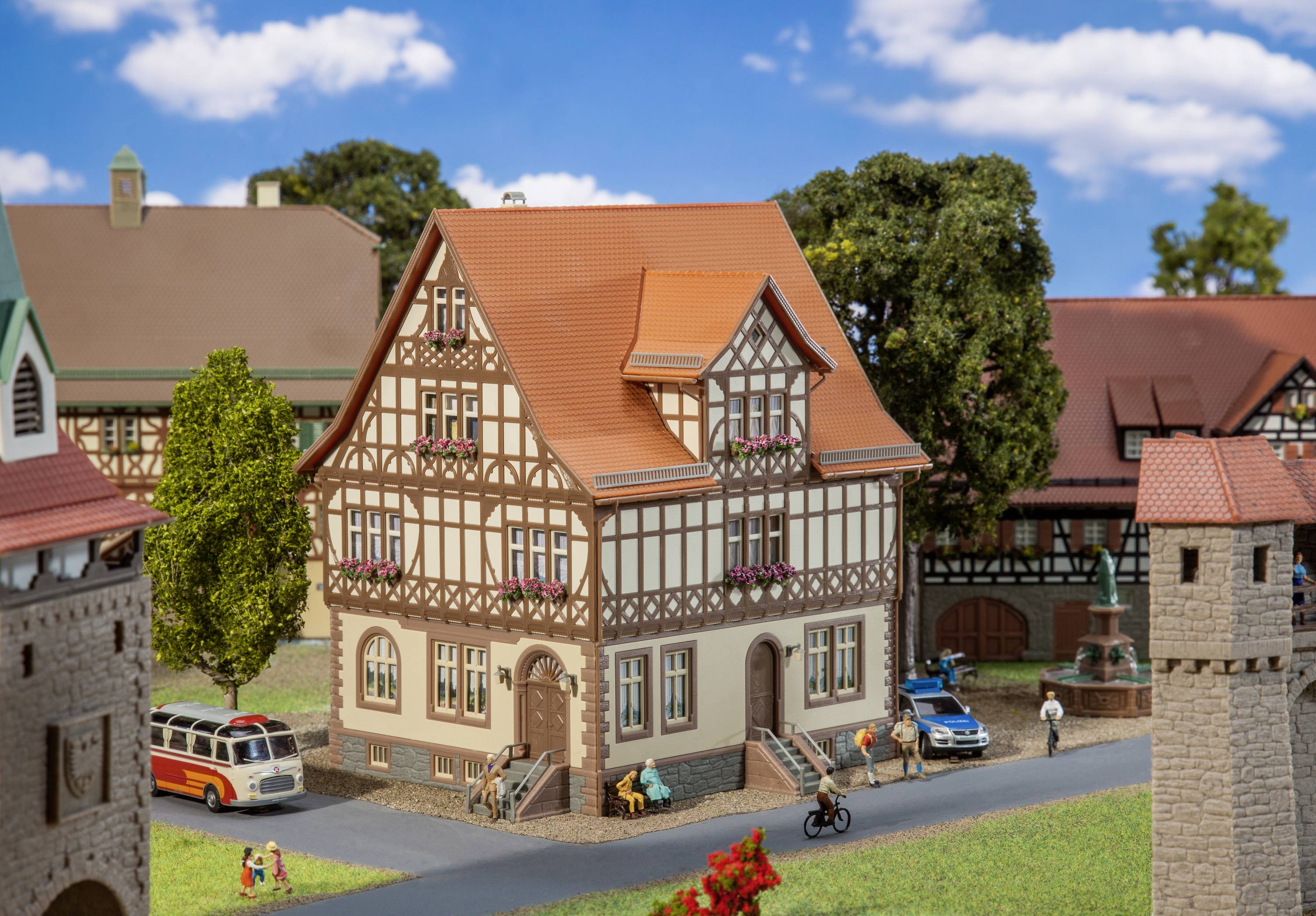 Un villaggio in miniatura mostra una casa a graticcio con tegole rosse, circondato da alberi, persone e veicoli in una scena idilliaca.