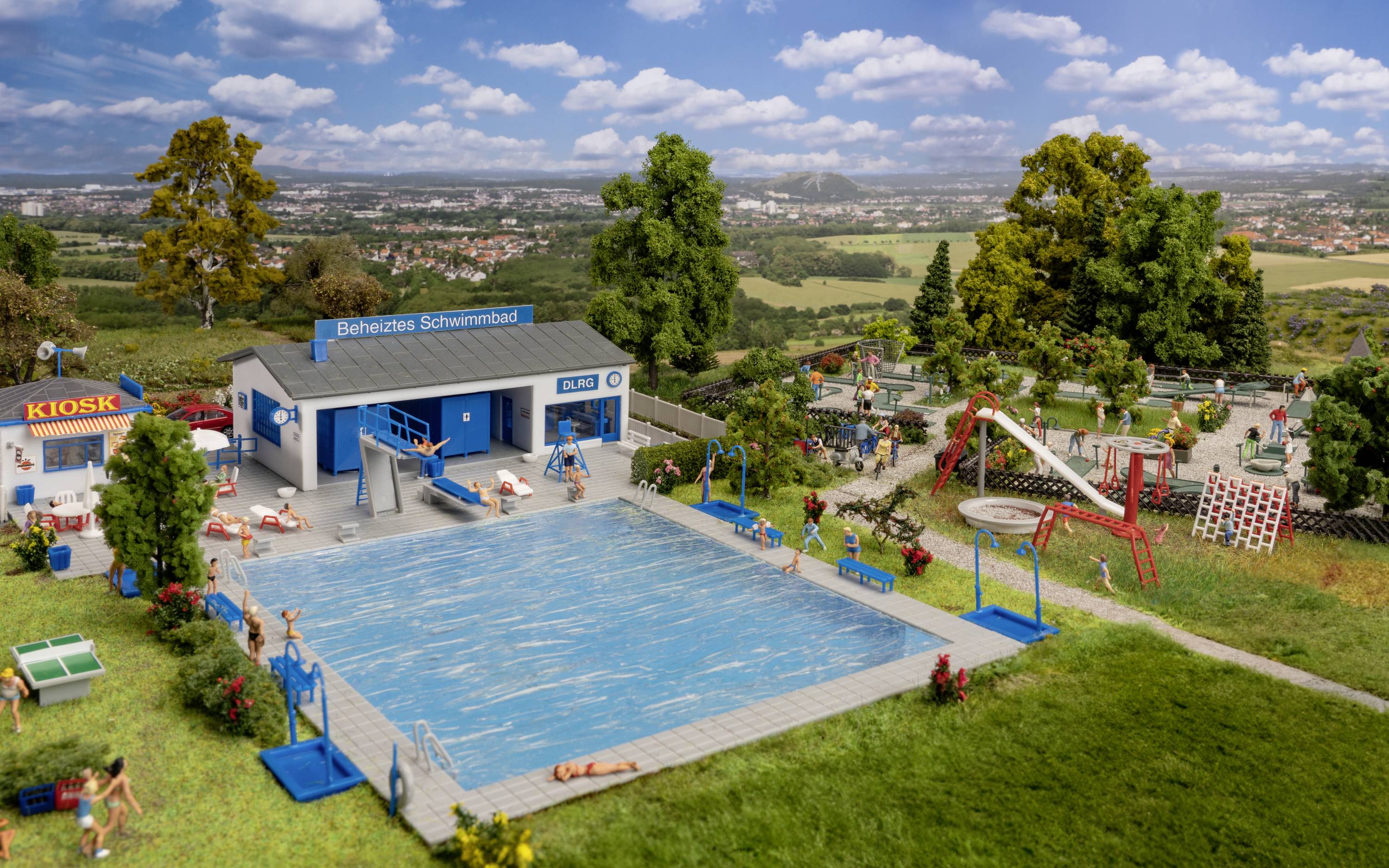 Un modello di piscina all'aperto con piscina riscaldata, chiosco e area giochi, circondato da alberi e prati in un ambiente rurale.