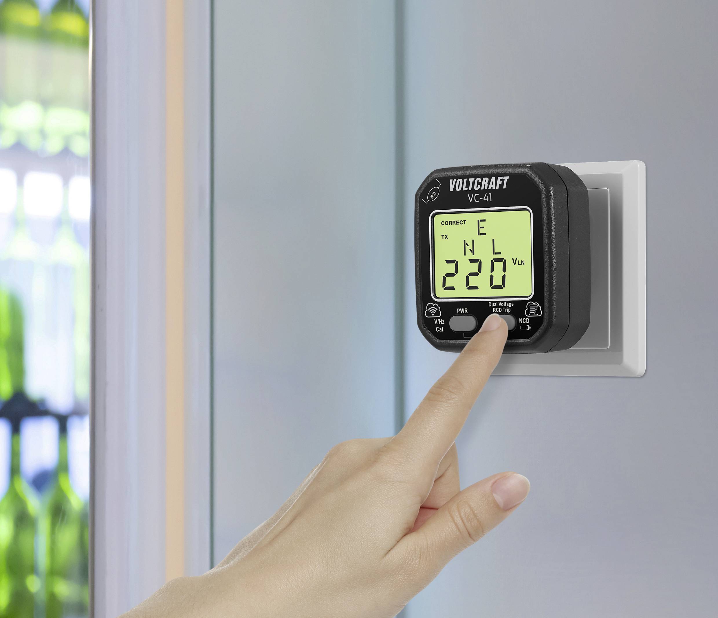 Una mano preme su uno strumento di misurazione elettrico con display '220 V'. Lo strumento è collegato a una presa a muro.