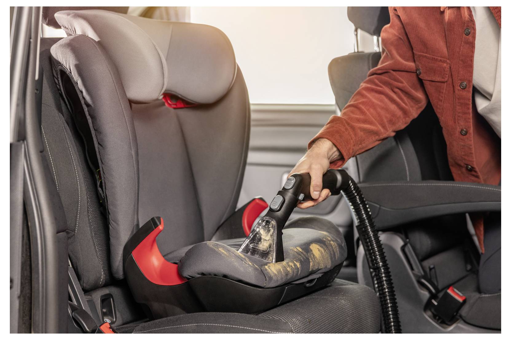 Una persona con una giacca rossa sta pulendo un seggiolino per bambini con un aspirapolvere, rimuovendo briciole e detriti all'interno dell'auto.