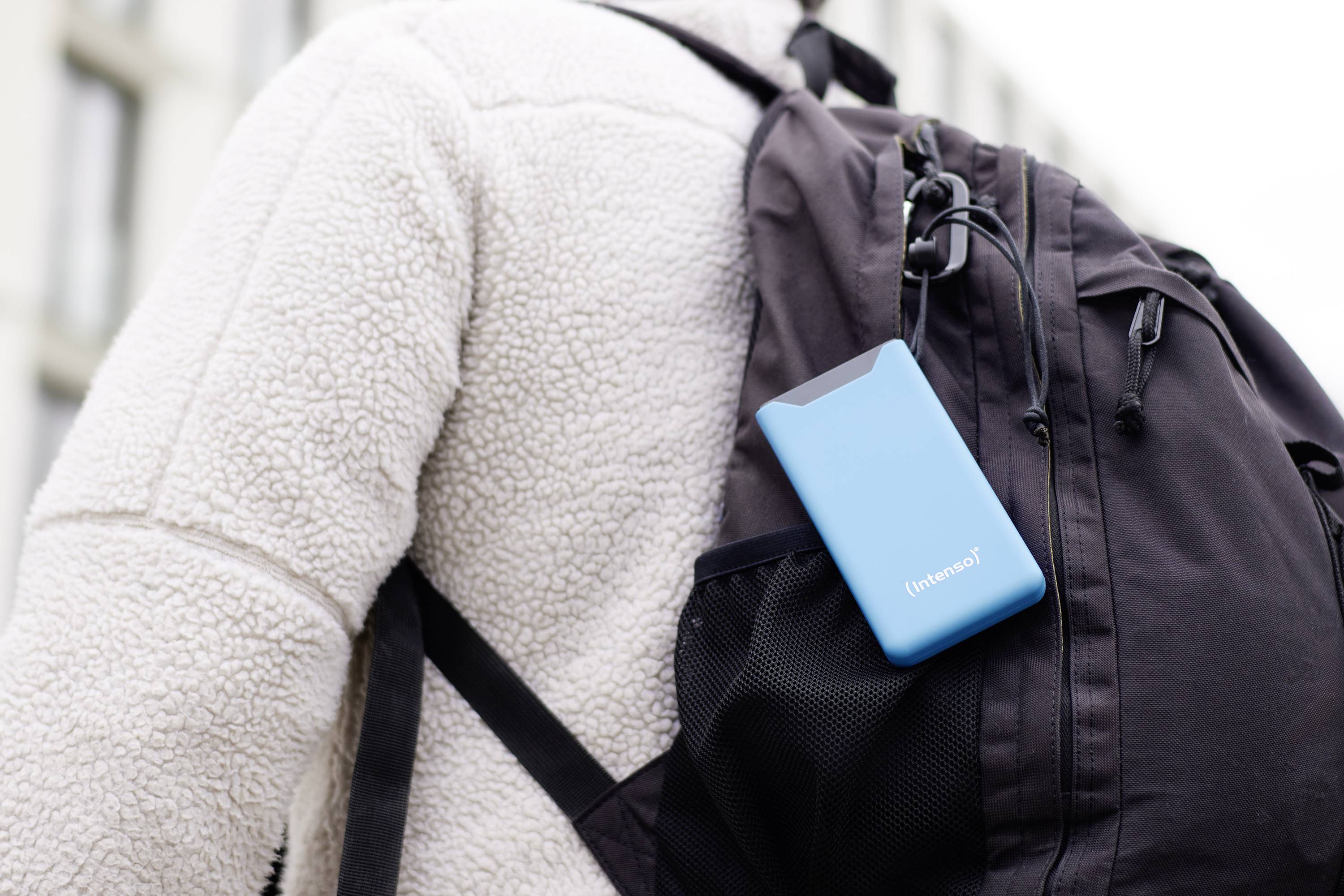 Una persona indossa uno zaino nero con un hard disk portatile blu nel tasca laterale. Edifici sono visibili sullo sfondo.