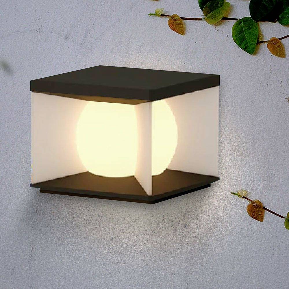 Applique con design cubico moderno che emana una luce calda. Circondato da alcune foglie su una parete chiara.
