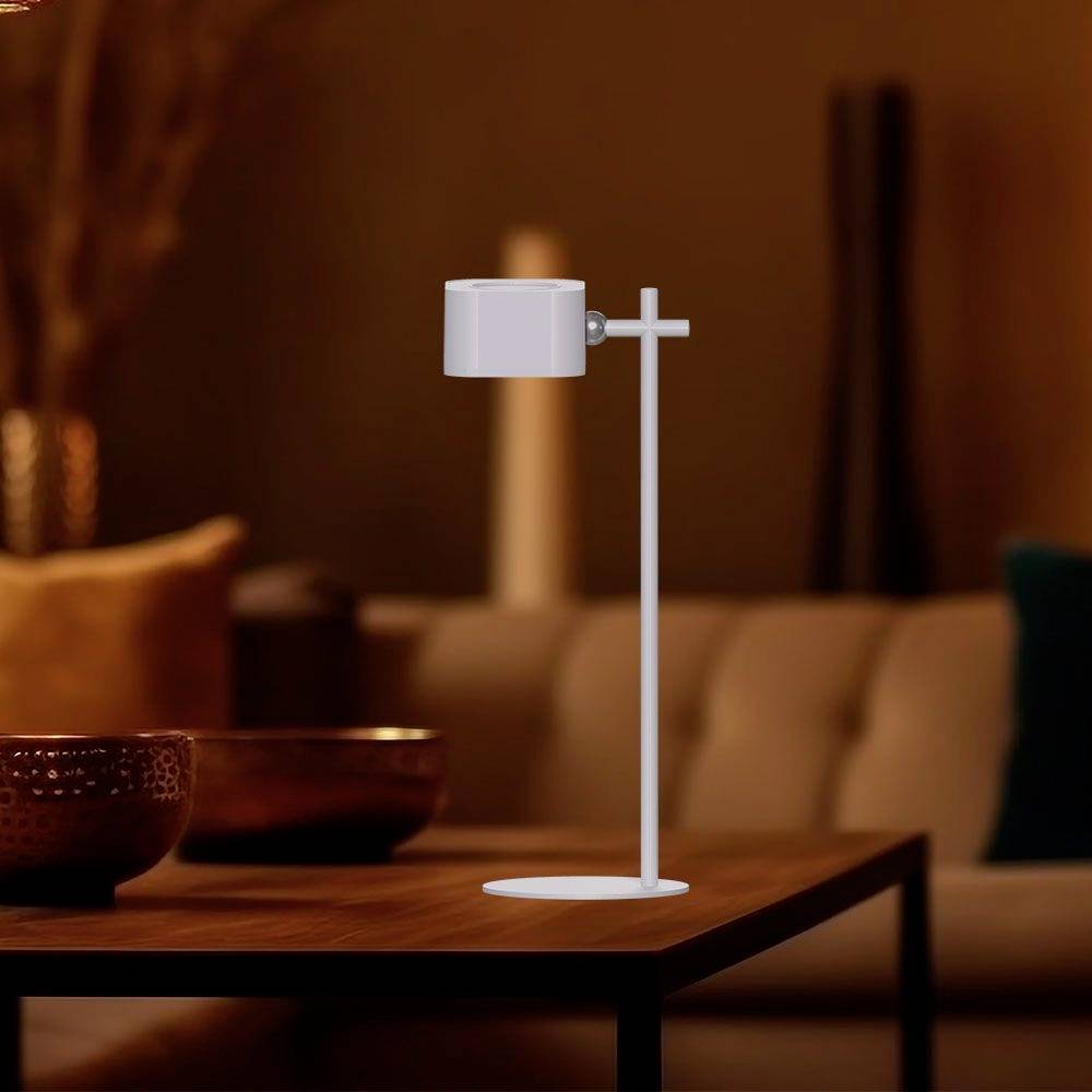 Una moderna lampada da tavolo LED bianca è posizionata su un tavolo di legno in un soggiorno accogliente, con un divano sullo sfondo.