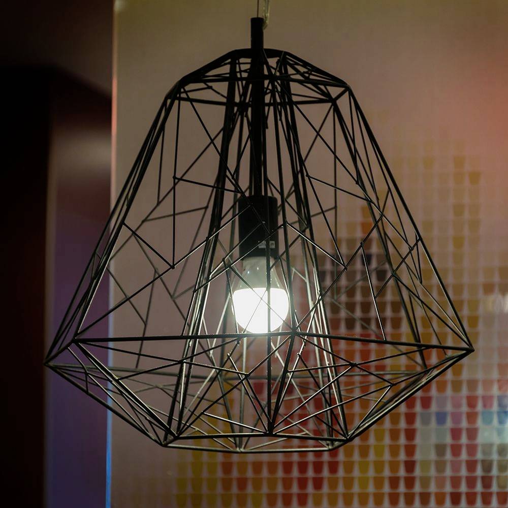 Un lampadario a sospensione nero a forma geometrica con elemento illuminante acceso, davanti a una parete colorata e decorata.