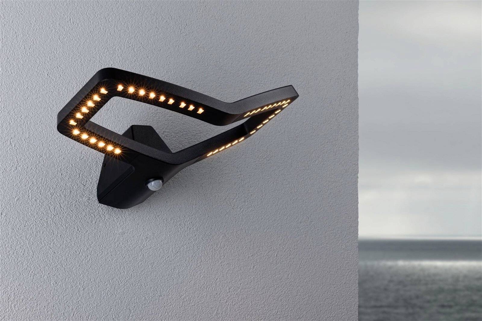 Una moderna lampada esterna nera con luci LED gialle è montata su una parete grigia. Sullo sfondo si vede il mare.