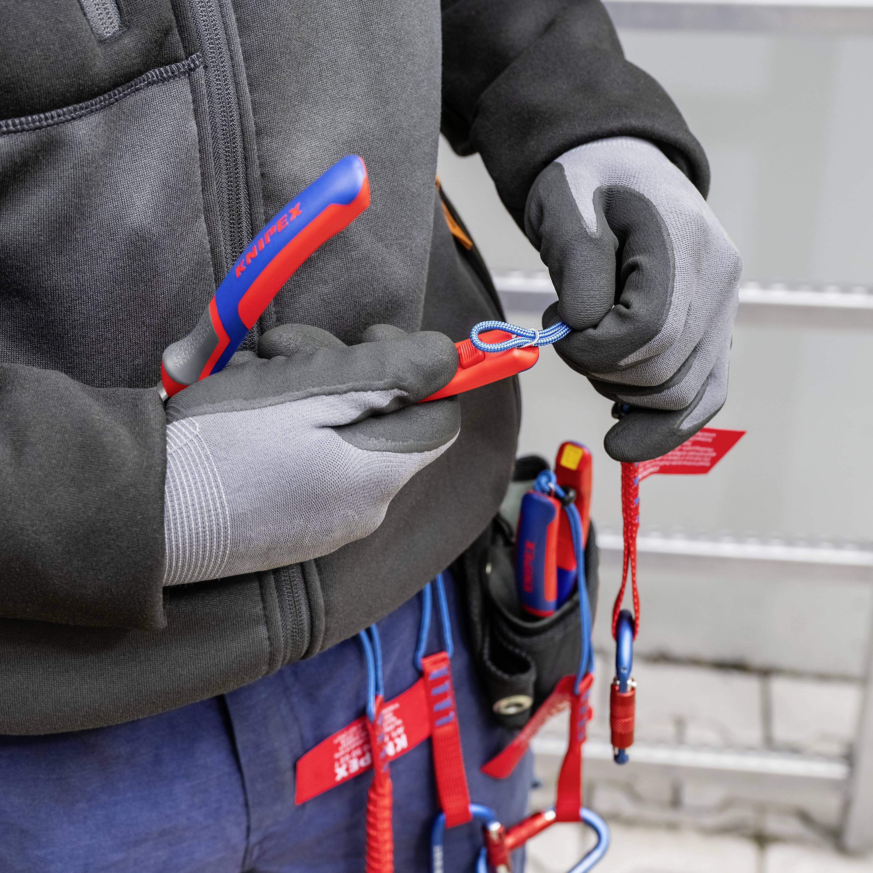 Una persona tiene un moschettone rosso e blu mentre indossa guanti da lavoro. L'attrezzatura da arrampicata è fissata alla sua cintura.