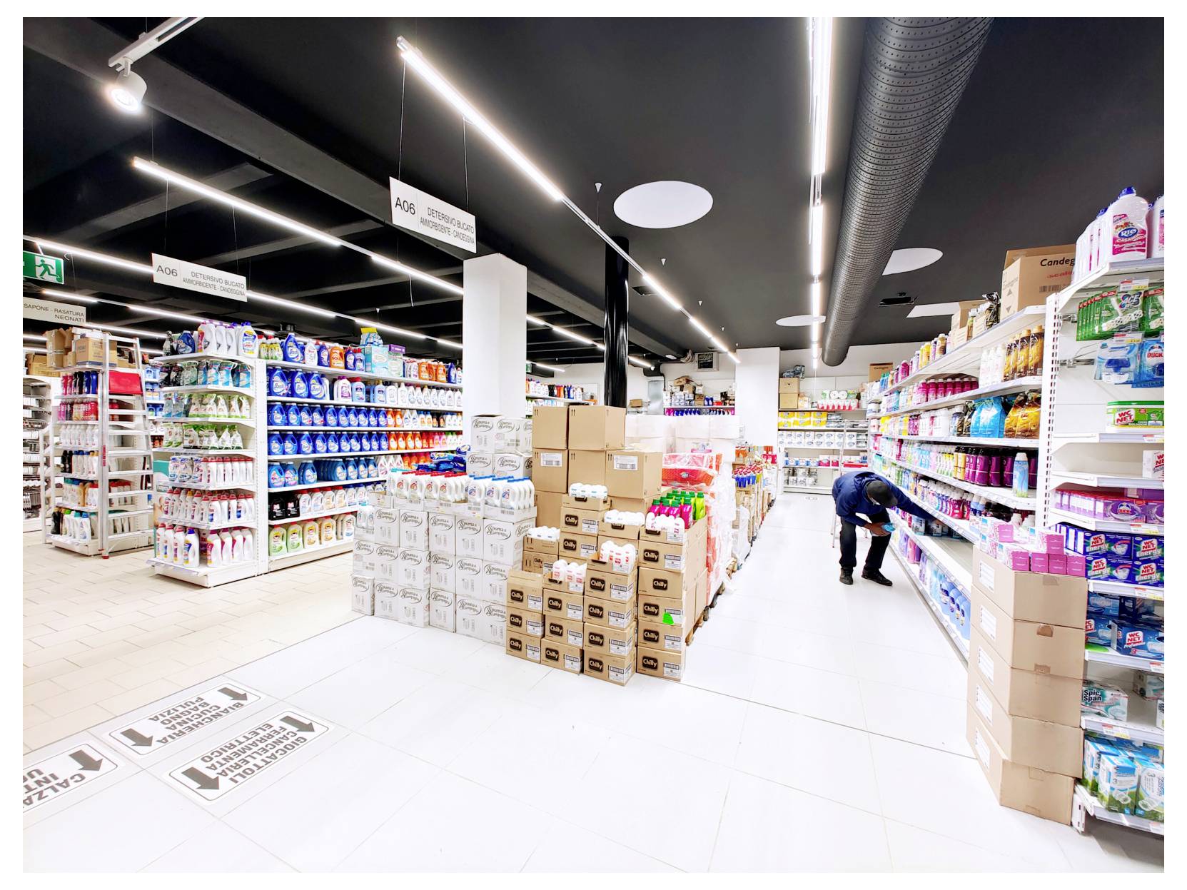Corsia luminosa di un supermercato con scaffali di generi alimentari e scatoloni di cartone impilati; un addetto rifornisce i prodotti sulla destra.