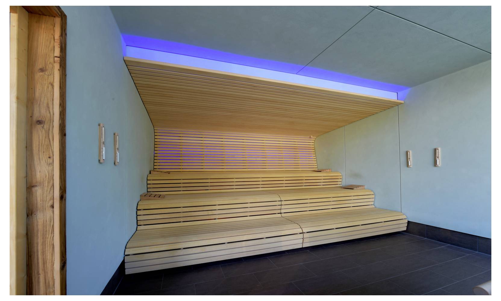 Stanza vuota e moderna con gradinate o piattaforma in legno contro una parete a listelli, illuminata da luci blu sul soffitto e pareti chiare.