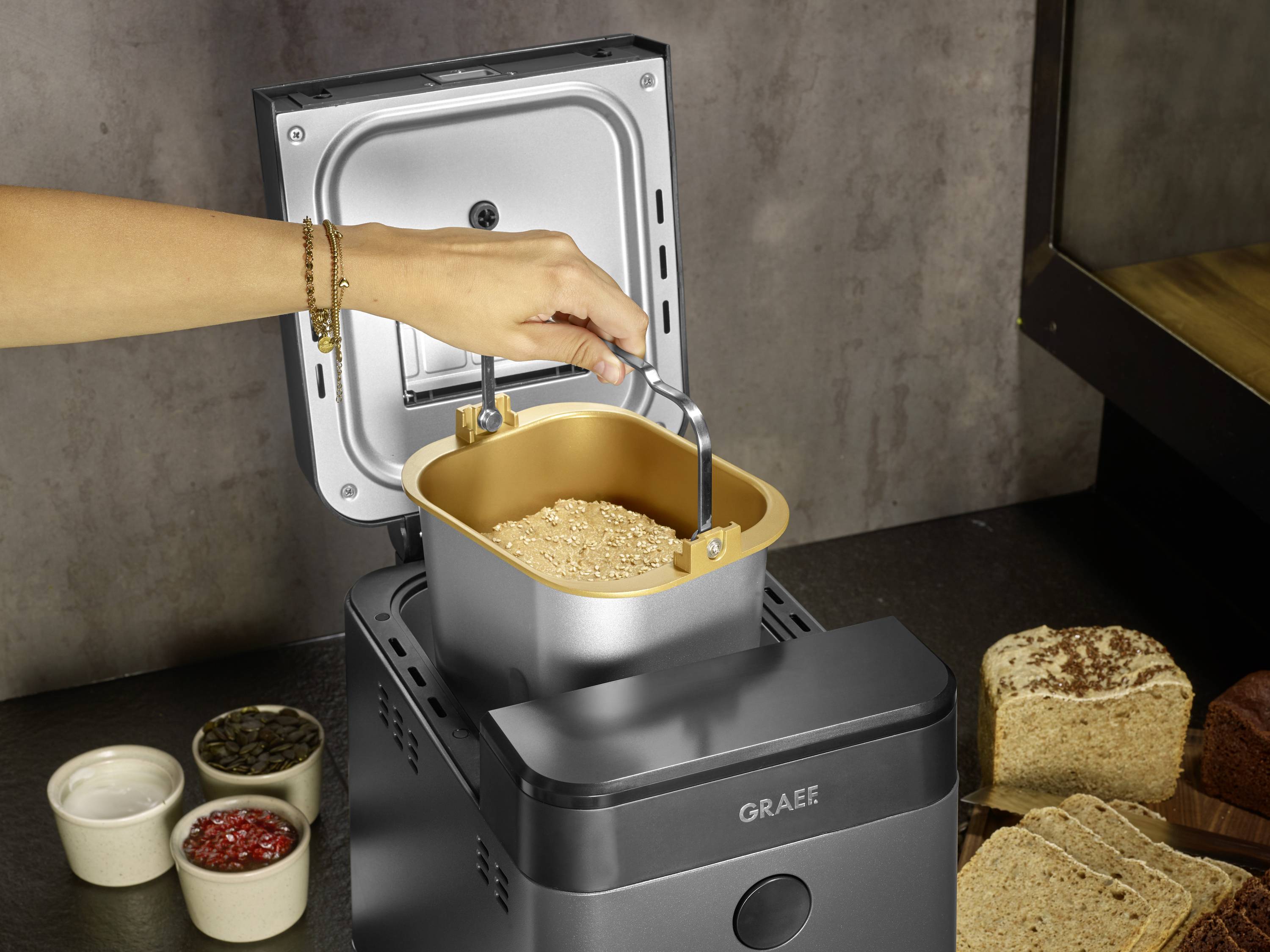 Una mano regge un contenitore per impastare con dell'impasto sopra un macchina per il pane aperta. Accanto ci sono pane e ingredienti.