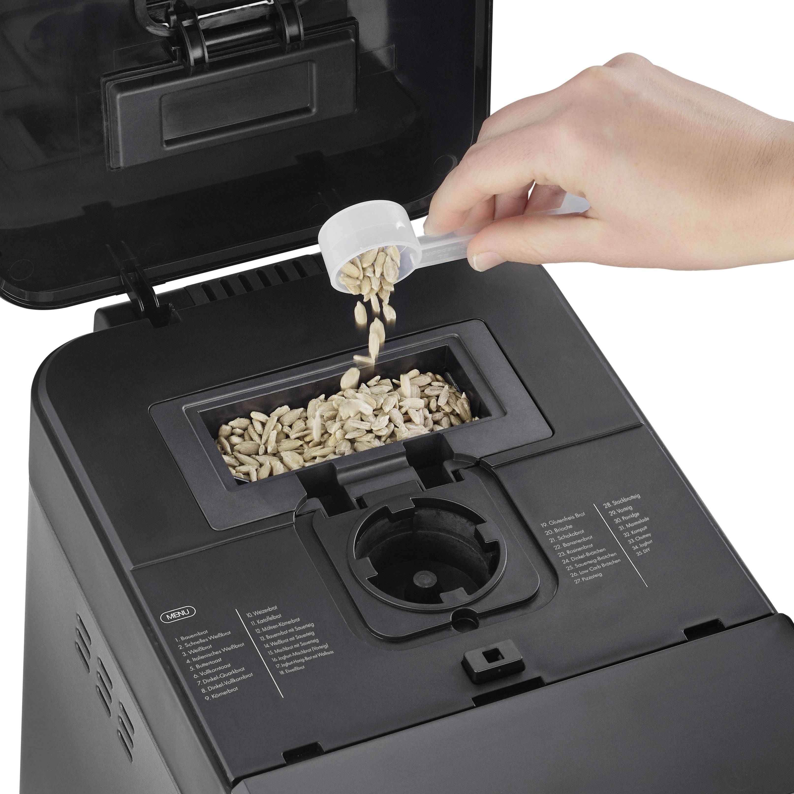 Una mano riempie semi di girasole in un misurino, versandoli nell'apertura superiore di una macchina per il pane nera.