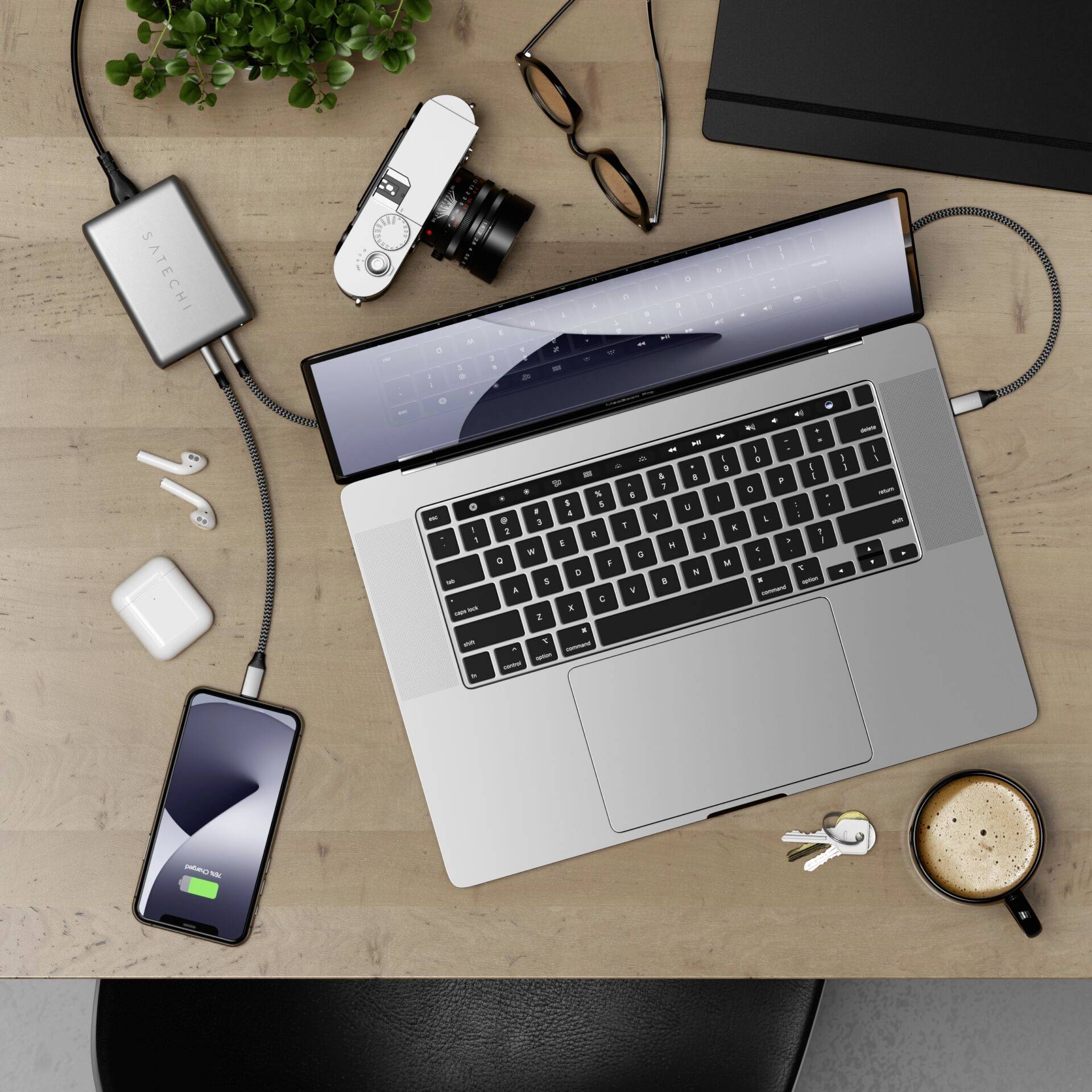 Una scrivania con laptop, smartphone, macchina fotografica, tazza di caffè, chiavi e piante. Lo smartphone è collegato a un caricabatterie.