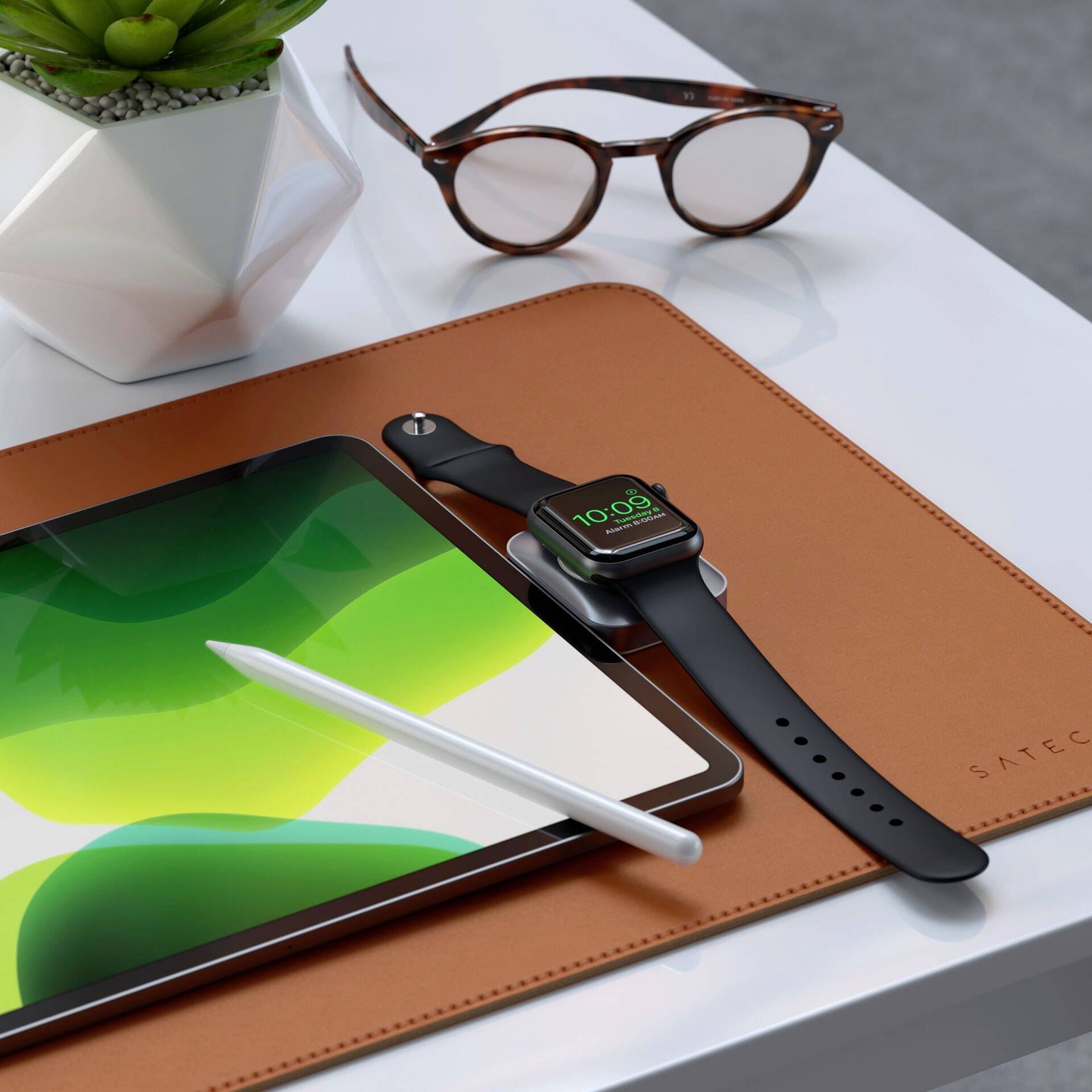 Tablet con penna su tappetino in pelle, accanto a un orologio intelligente con quadrante verde; sullo sfondo occhiali e una pianta in un vaso geometrico.