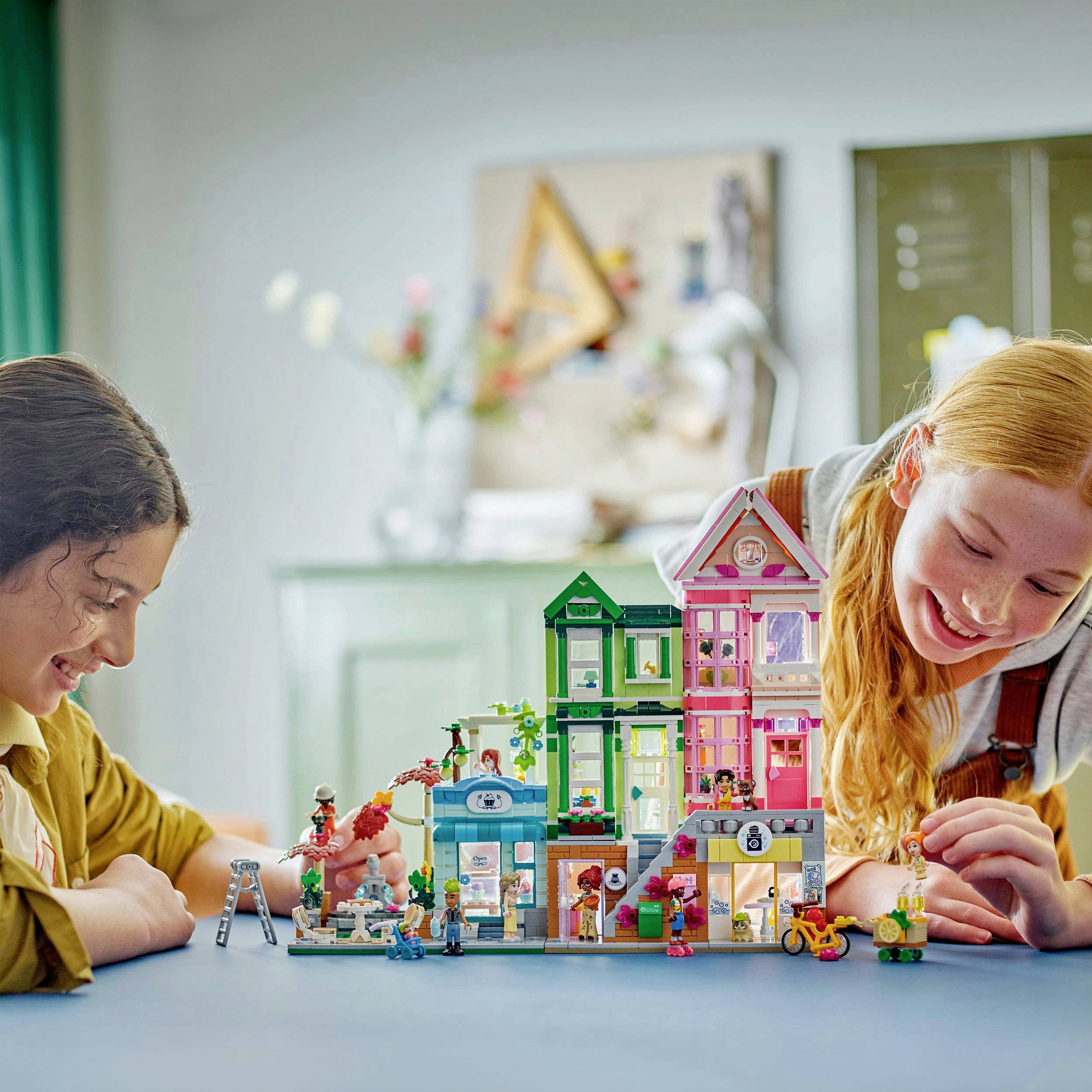 Due bambini giocano con un modello dettagliato e colorato di una casa cittadina costruito con blocchi. Entrambi sorridono felici.