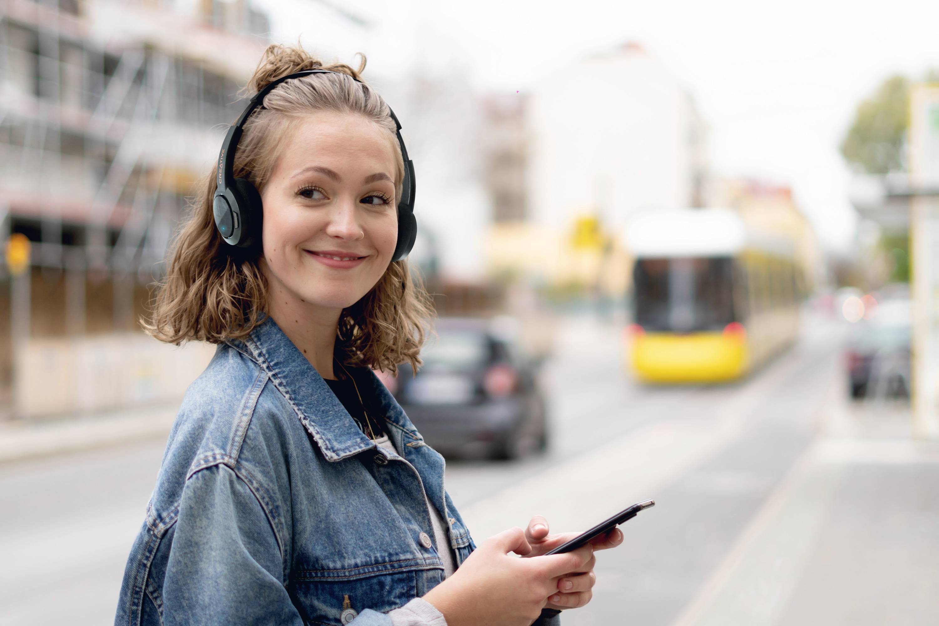 Una donna con le cuffie e lo smartphone sorride ai bordi della strada. Sullo sfondo, un tram giallo passa accanto a lei.