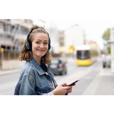 Una donna con le cuffie e lo smartphone sorride ai bordi della strada. Sullo sfondo, un tram giallo passa accanto a lei.