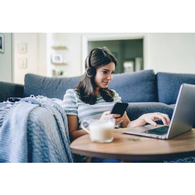 Una donna è seduta sul divano, indossa delle cuffie e lavora al laptop, mentre tiene in mano uno smartphone. In primo piano c'è un tavolo con una bevanda.