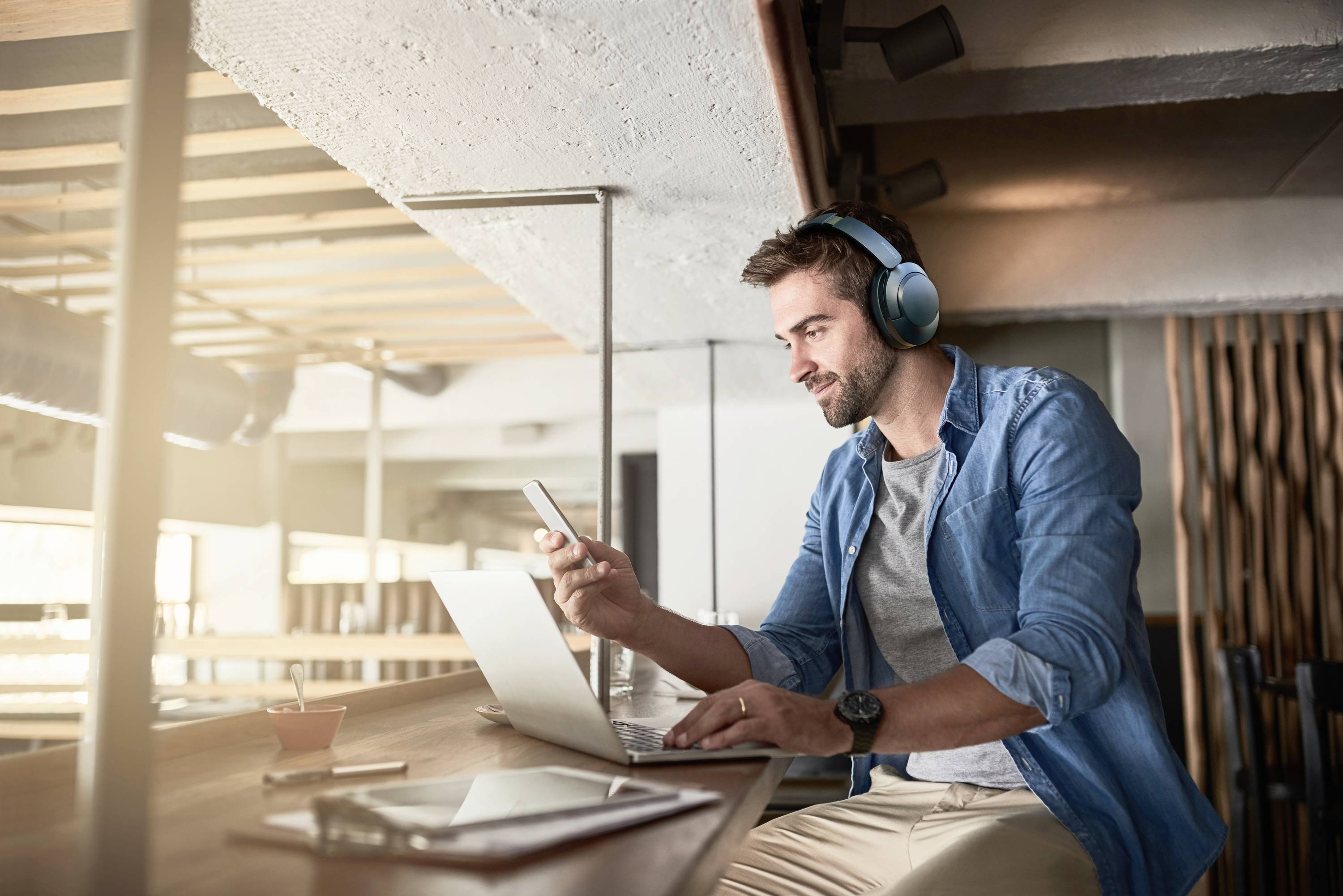 Un uomo con le cuffie lavora in un caffè moderno su un laptop, utilizzando anche uno smartphone. Appare concentrato e rilassato.