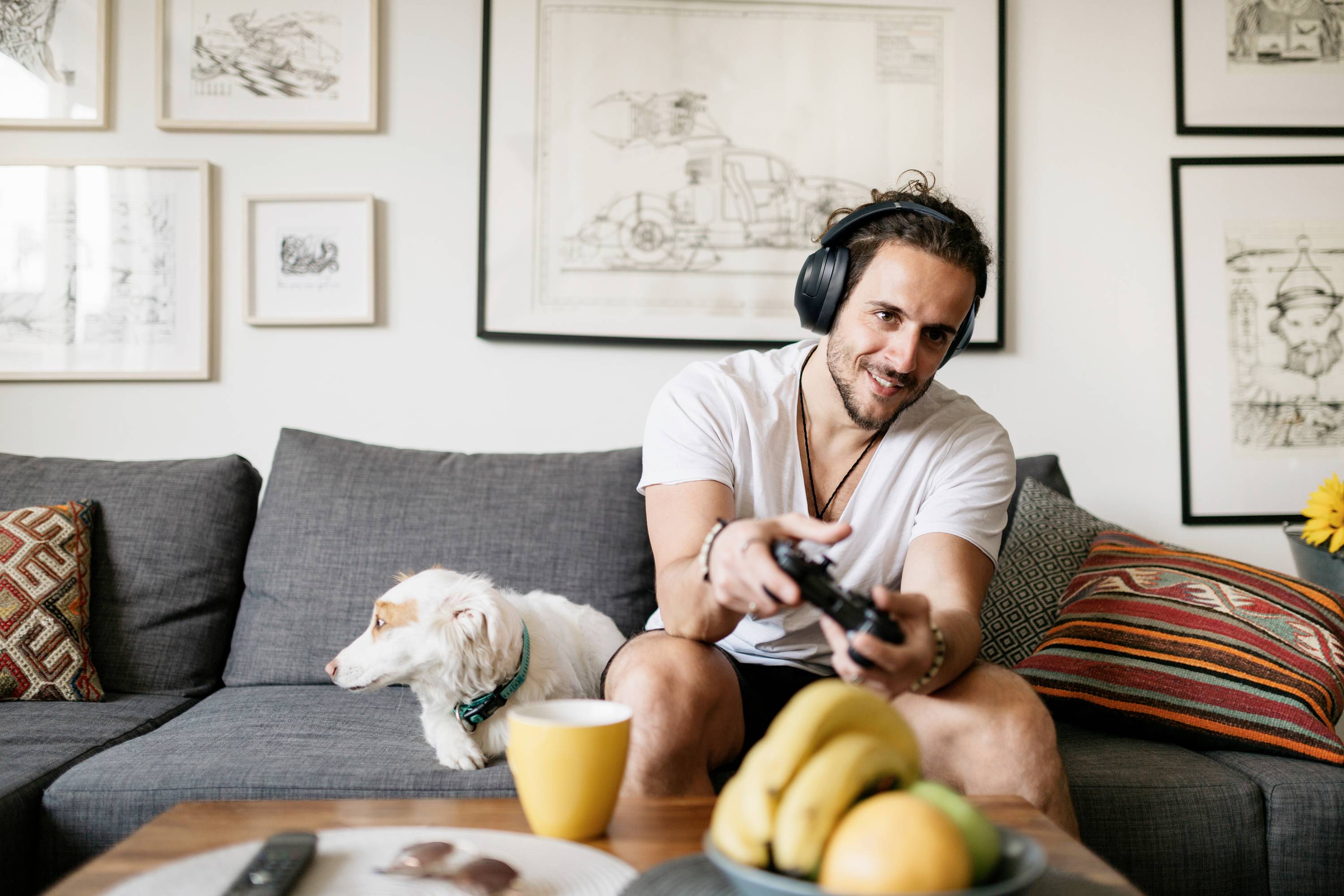 Uomo con le cuffie che gioca a un videogioco sul divano. Un cane gli siede accanto. Soggiorno sullo sfondo pieno di cornici con immagini.