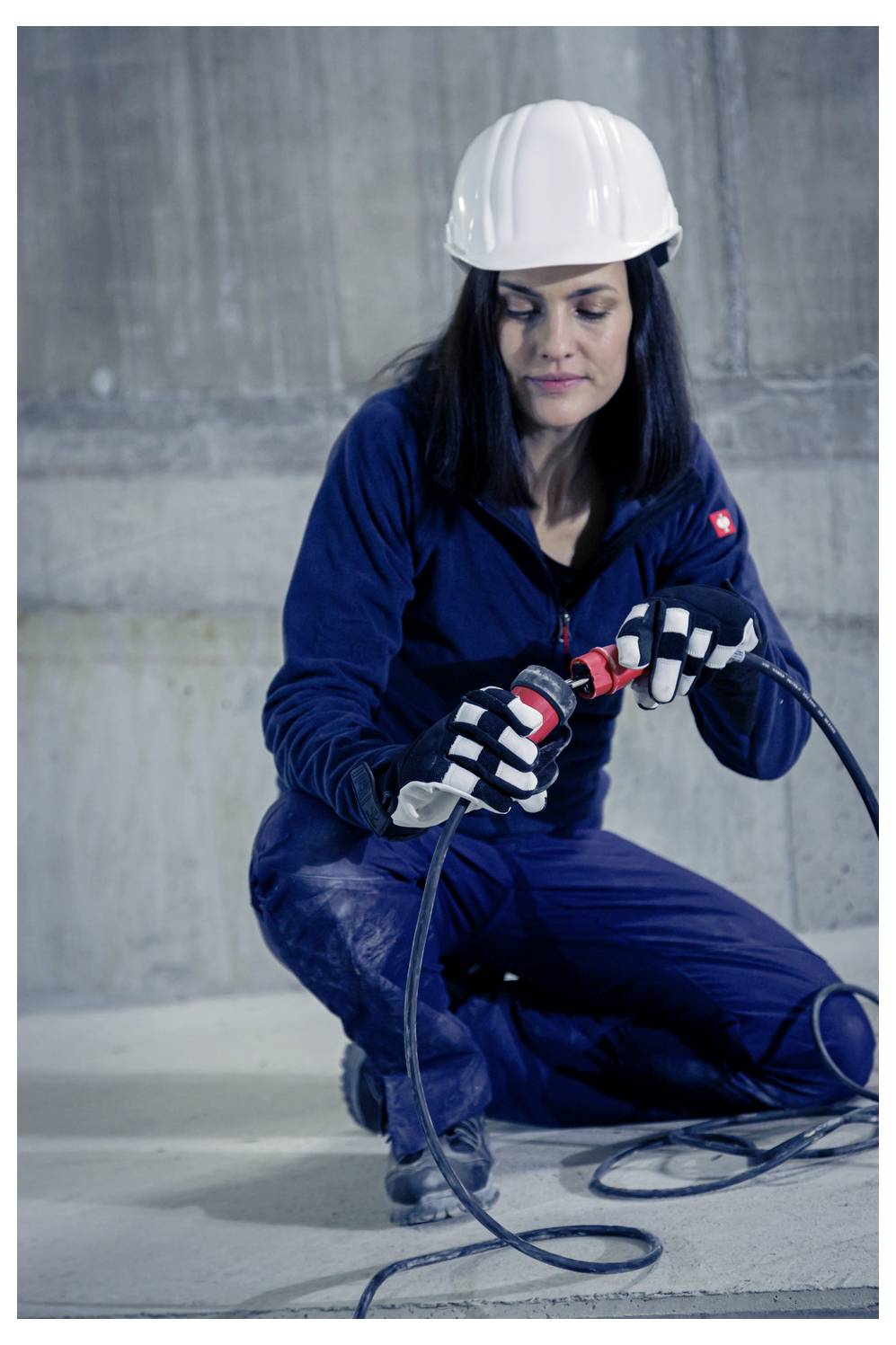Operaia edile in ginocchio su un pavimento in cemento, che utilizza un utensile elettrico con cavo mentre indossa un casco di sicurezza, guanti e una tuta da lavoro blu.