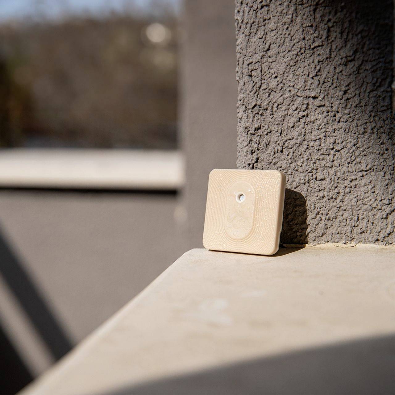 Un dispositivo beige e quadrato con un pulsante è poggiato su una superficie di cemento grigio sotto la luce del sole. Sullo sfondo, un paesaggio sfocato.